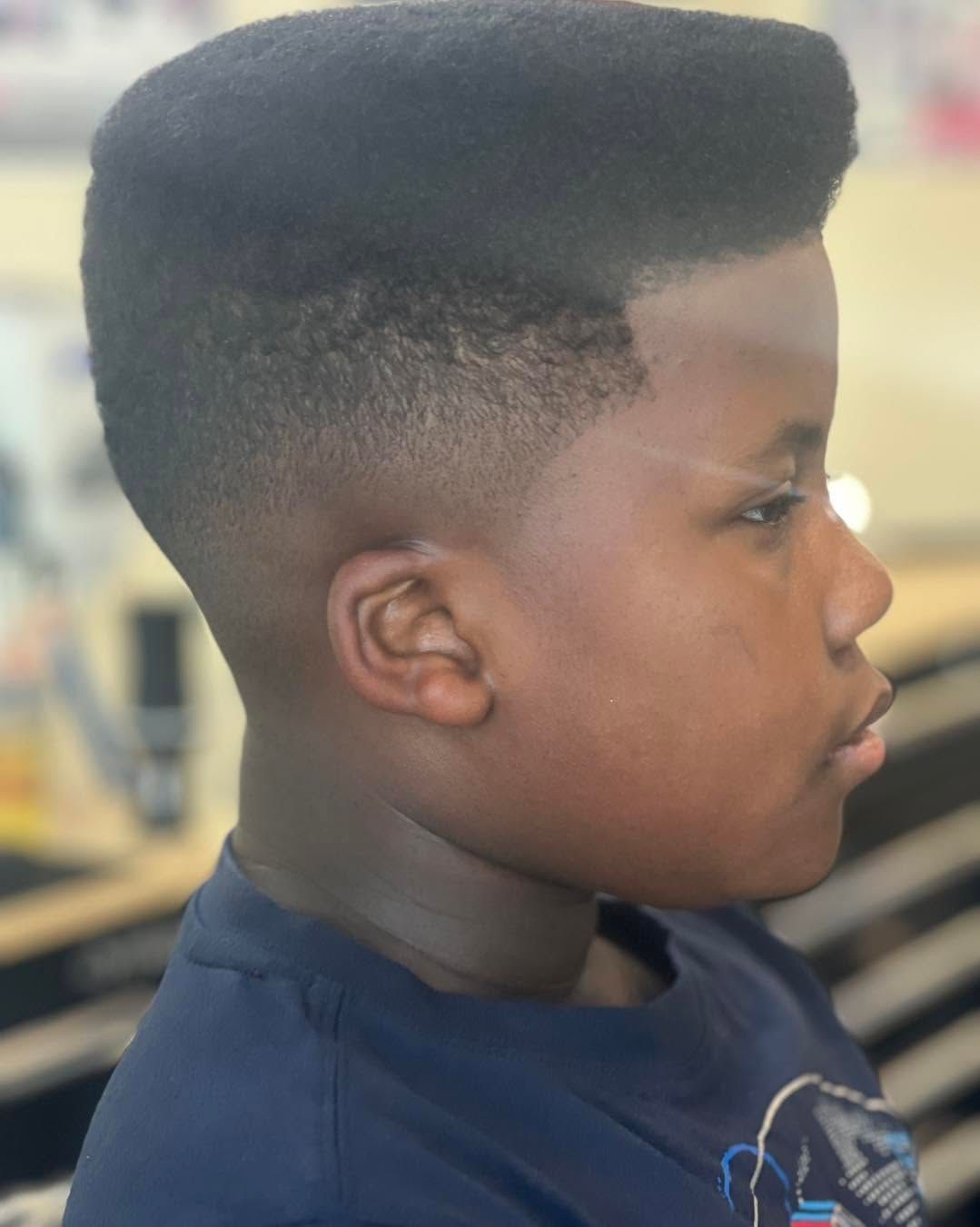 Profile of a child with a high flat-top haircut, wearing a dark shirt, in an indoor setting.