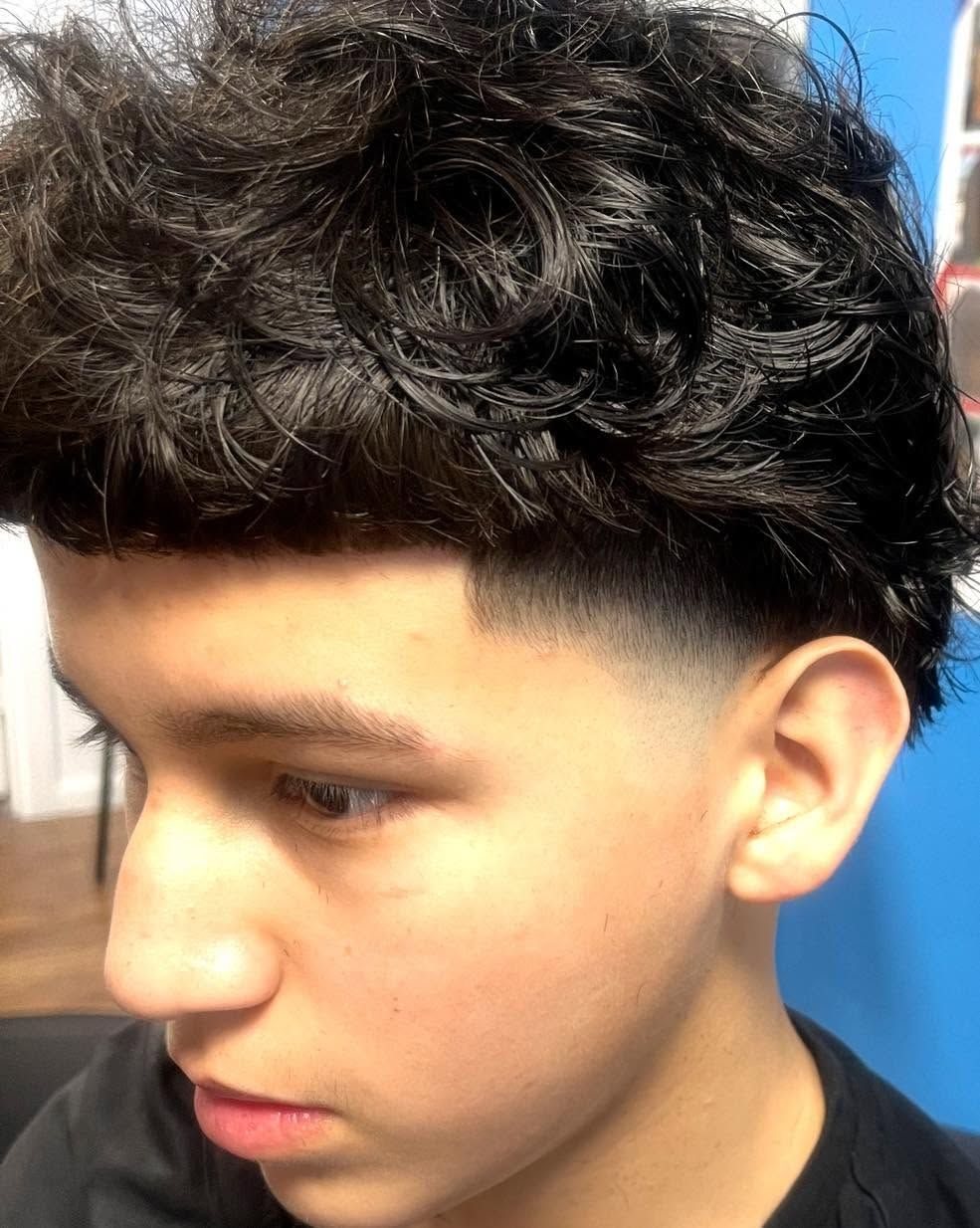 Side profile of a person with curly dark hair and a fade haircut against a blue background.