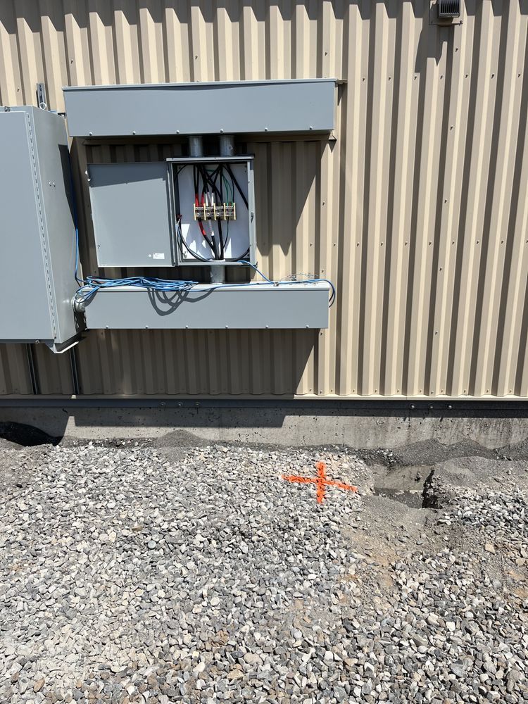 Electrical panel mounted on corrugated wall beside gravel ground, with orange utility marker nearby
