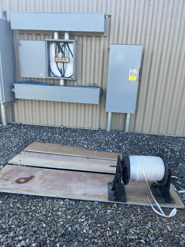 Electrical panels and conduit on a corrugated wall beside a spool of cable on gravel.