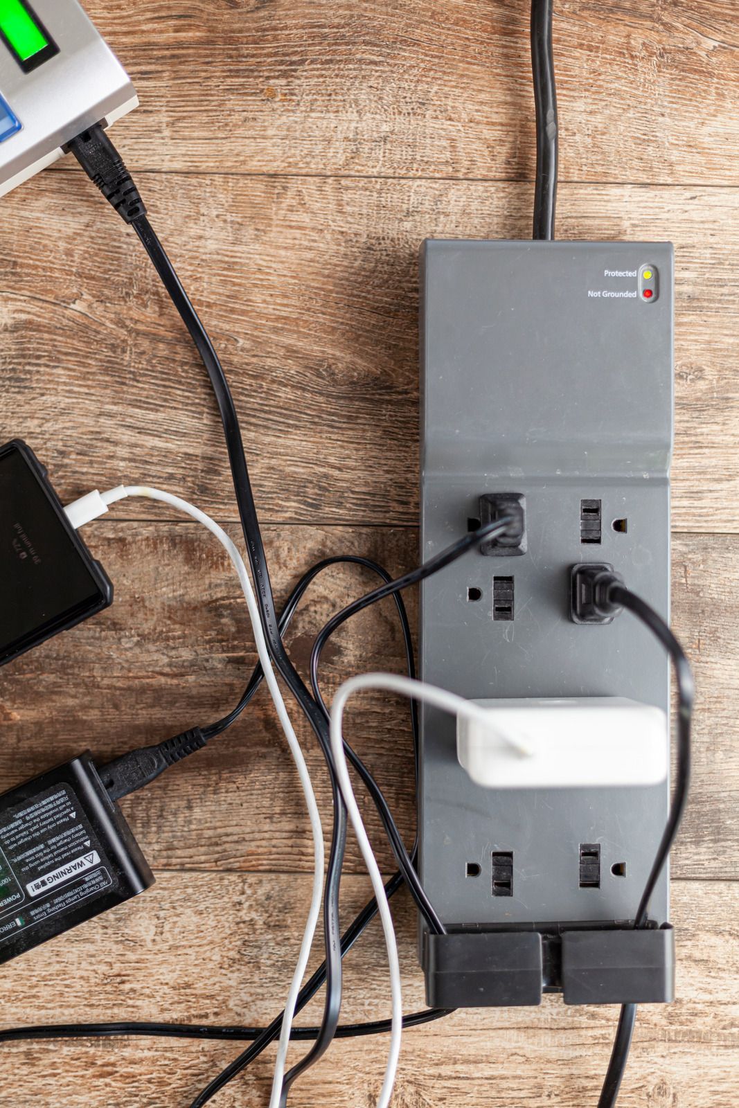 Power strip with multiple plugged-in cords on a wood floor, next to a black device and a white adapter