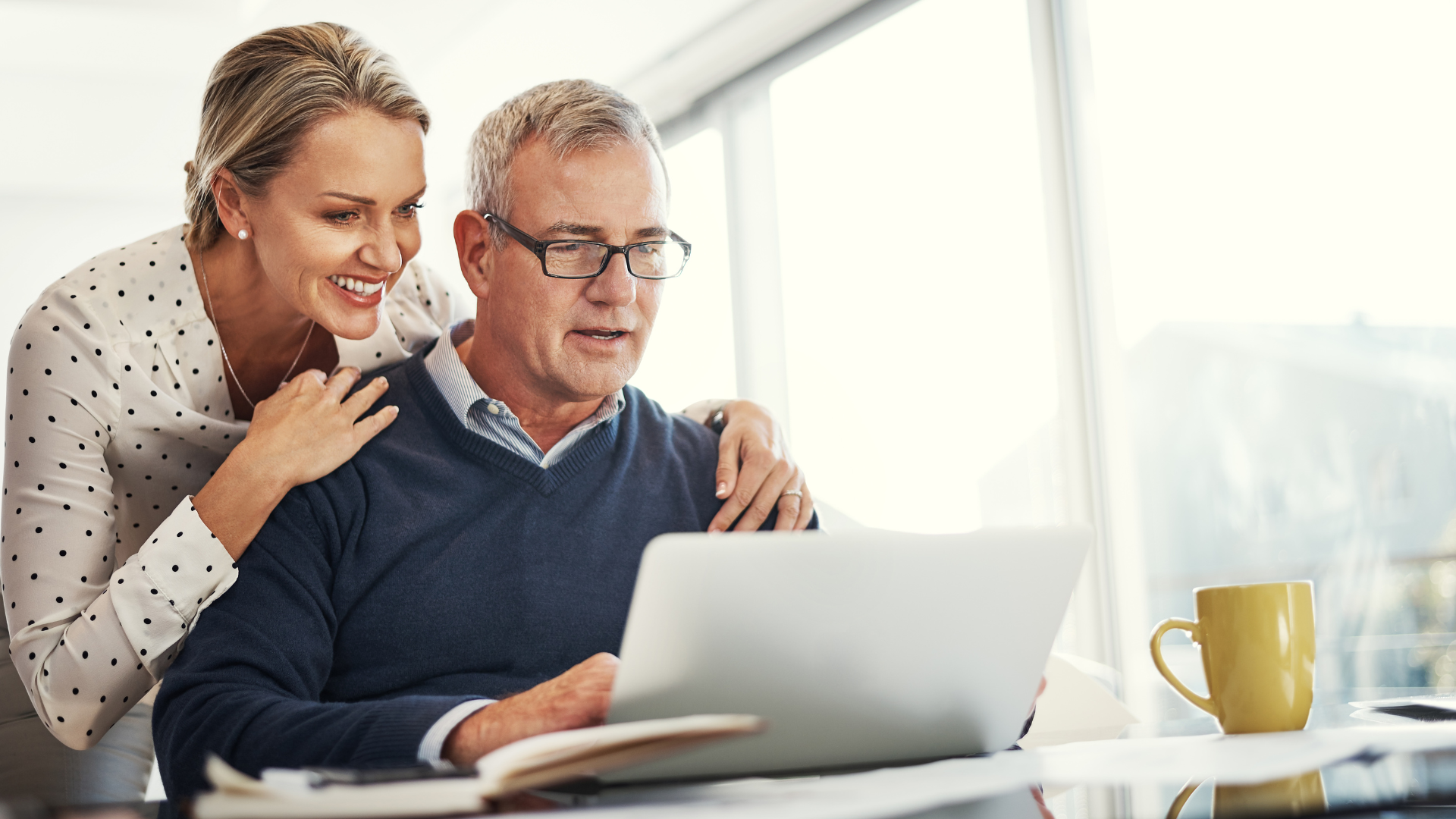 Woman smiles, puts arm around man looking at laptop; sunny room, yellow mug.