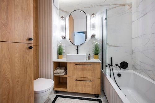 Bright modern bathroom with wood vanity, oval mirror, white tub, and marble tile walls.
