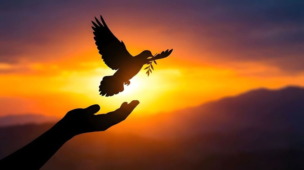 A silhouette of a hand releasing a dove holding an olive branch against a vibrant orange sunset over mountains.
