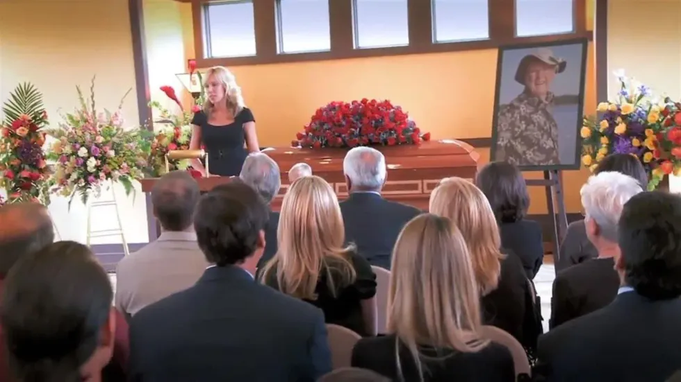 A woman speaks at a funeral service before a casket, floral arrangements, and a framed portrait of a person.