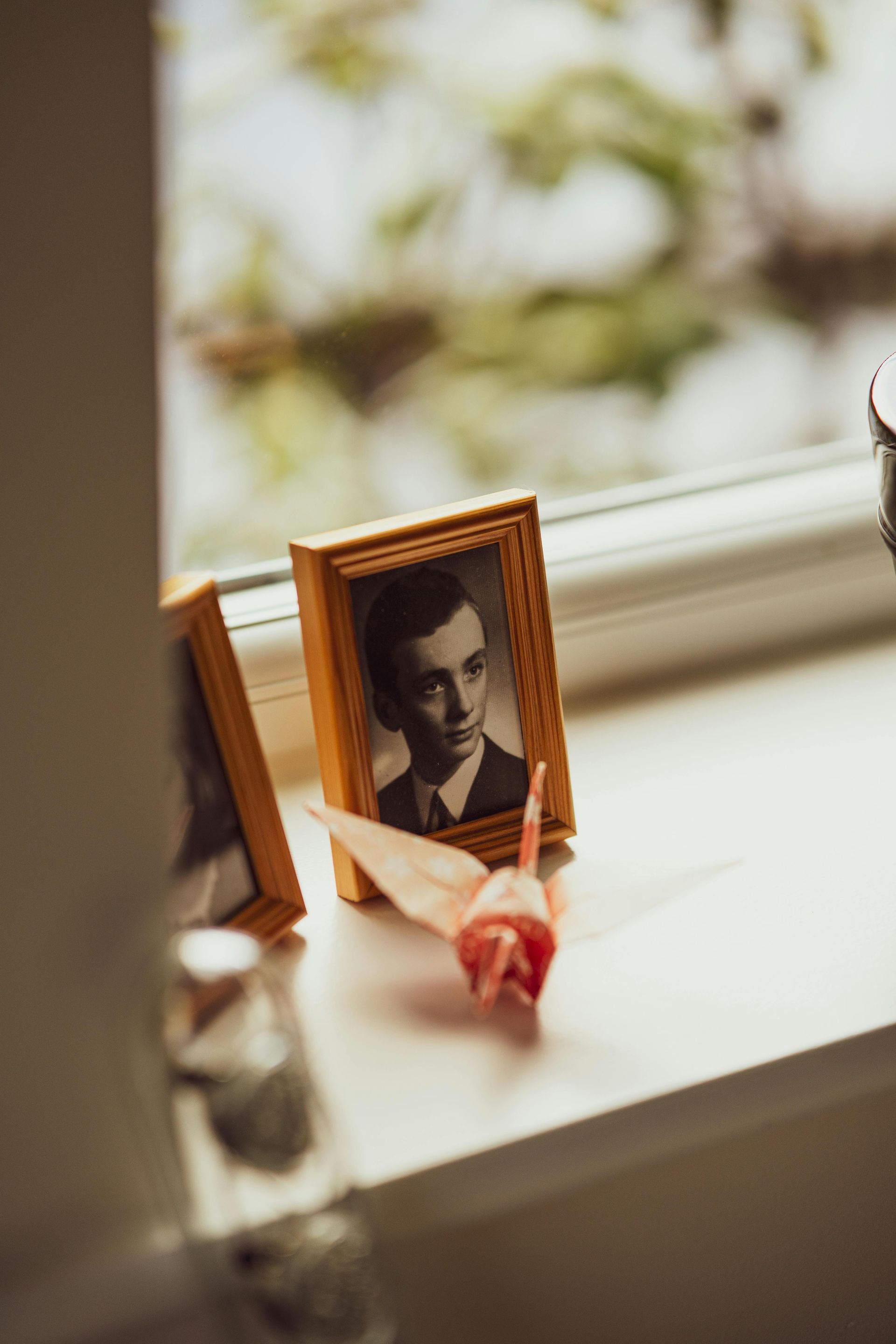 A vintage framed portrait of a person next to a folded paper crane on a sunlit windowsill.
