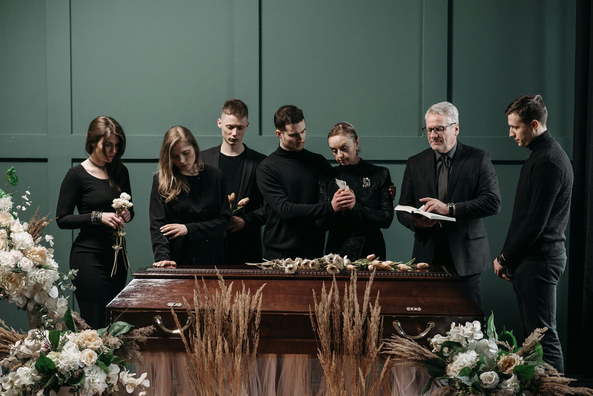 A group of people dressed in black stands solemnly around a wooden casket, with one person reading from an open book.