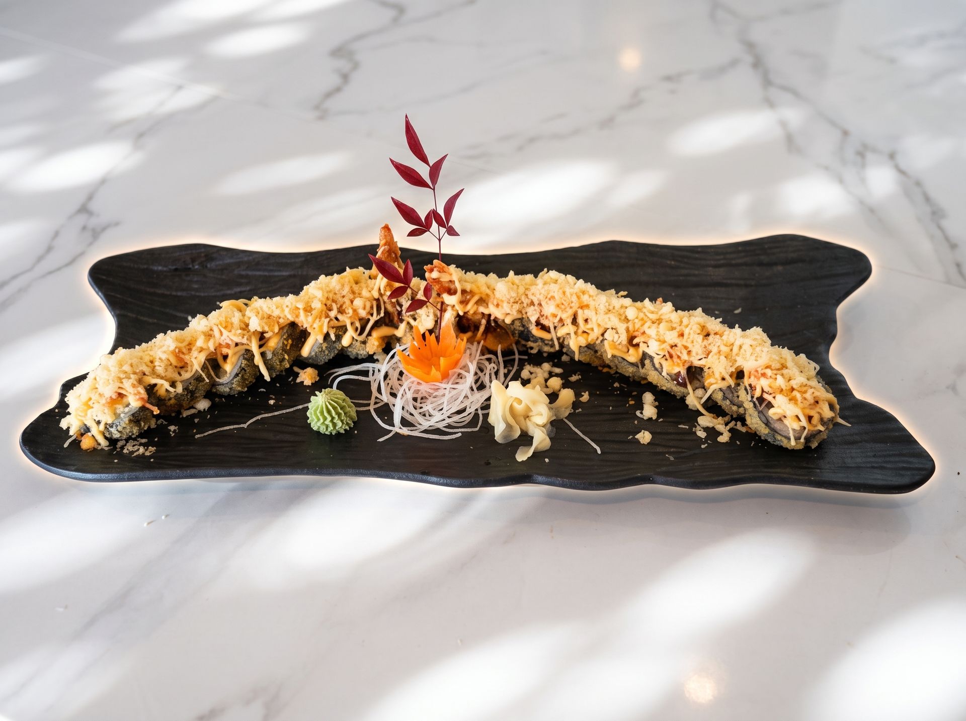 Sushi roll plated on a black rectangular dish, garnished with sauce and red herbs on a marble table