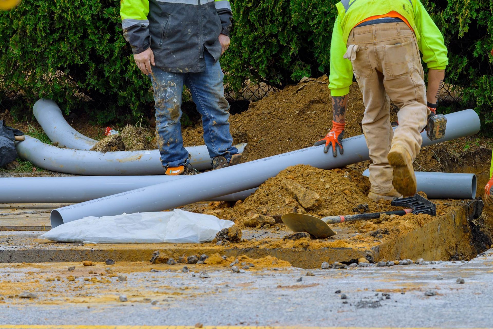 Construction workers near an open trench with gray pipes and dirt. One worker is walking.