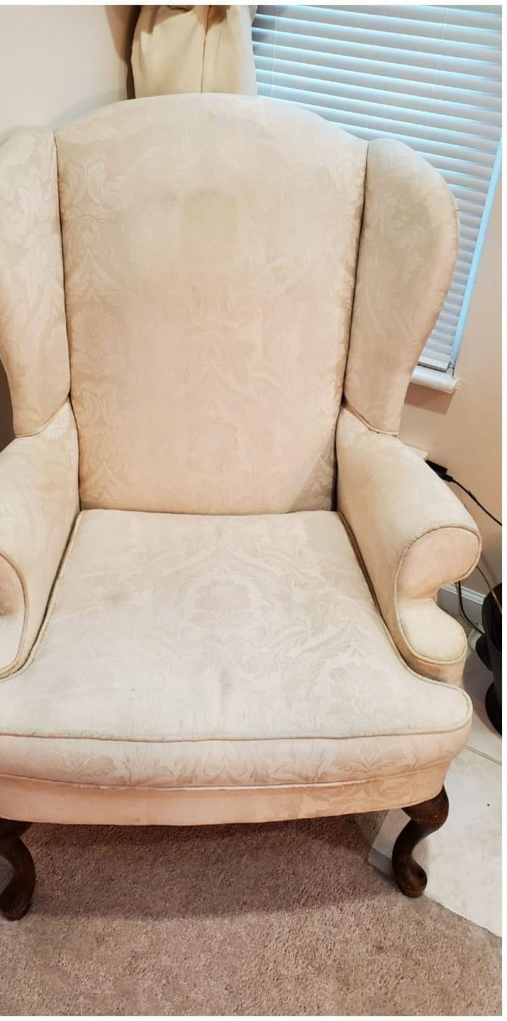 A cream-colored wingback chair with dark wooden legs sits on a brown carpet in front of a window.