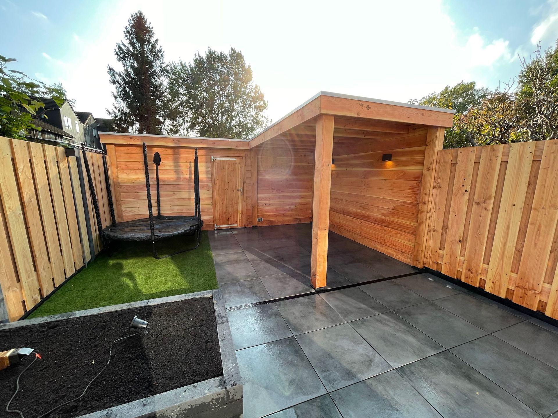 Achtertuin met houten schuur, trampoline en grijs terras. Aan beide zijden staan ​​hekken, met groen gras en een zonnige hemel.