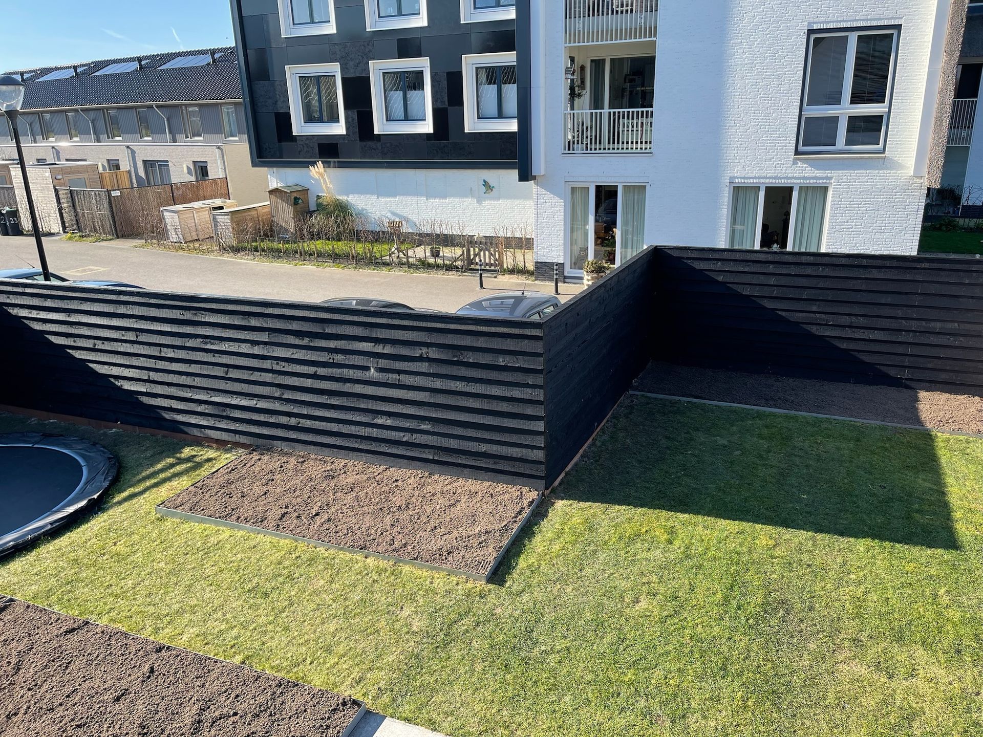 Een zwart houten hek omringt een grasveld met een trampoline voor een modern gebouw.