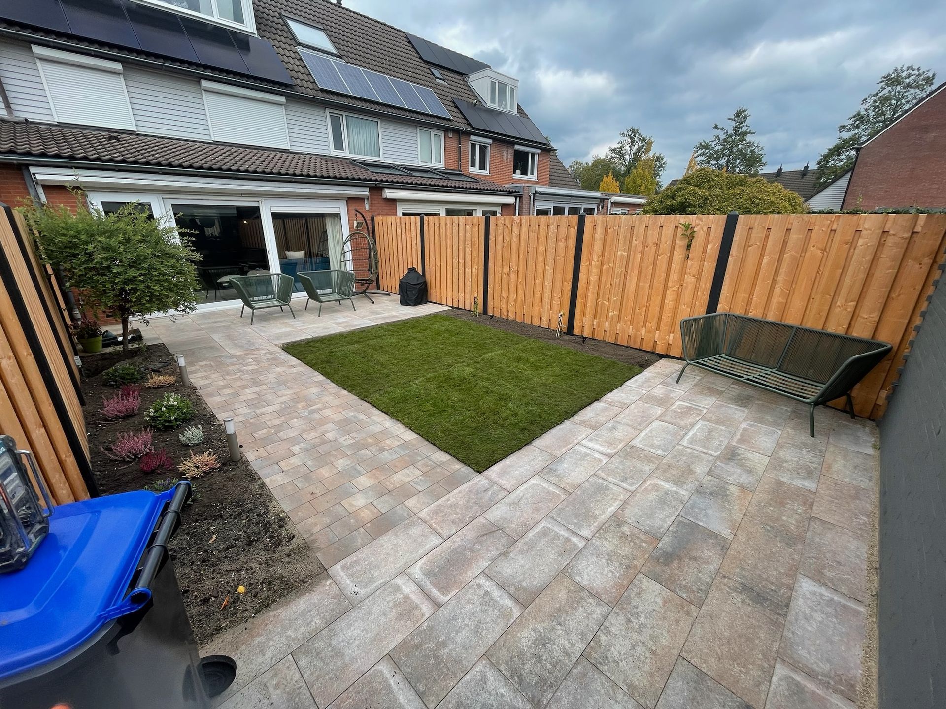 Een terras in de achtertuin met stenen bestrating, een klein gazon en een houten schutting.