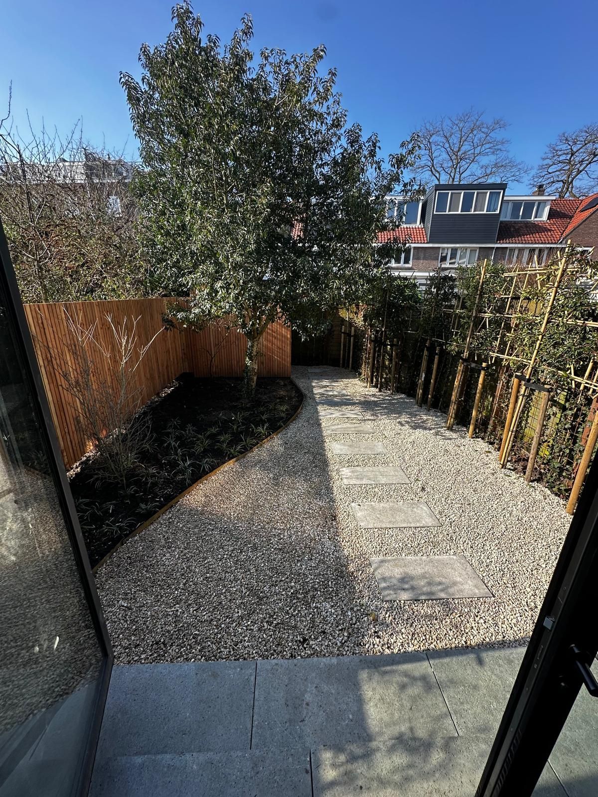 Achtertuin met stenen pad, grind, bamboehek en groen onder een helderblauwe hemel.