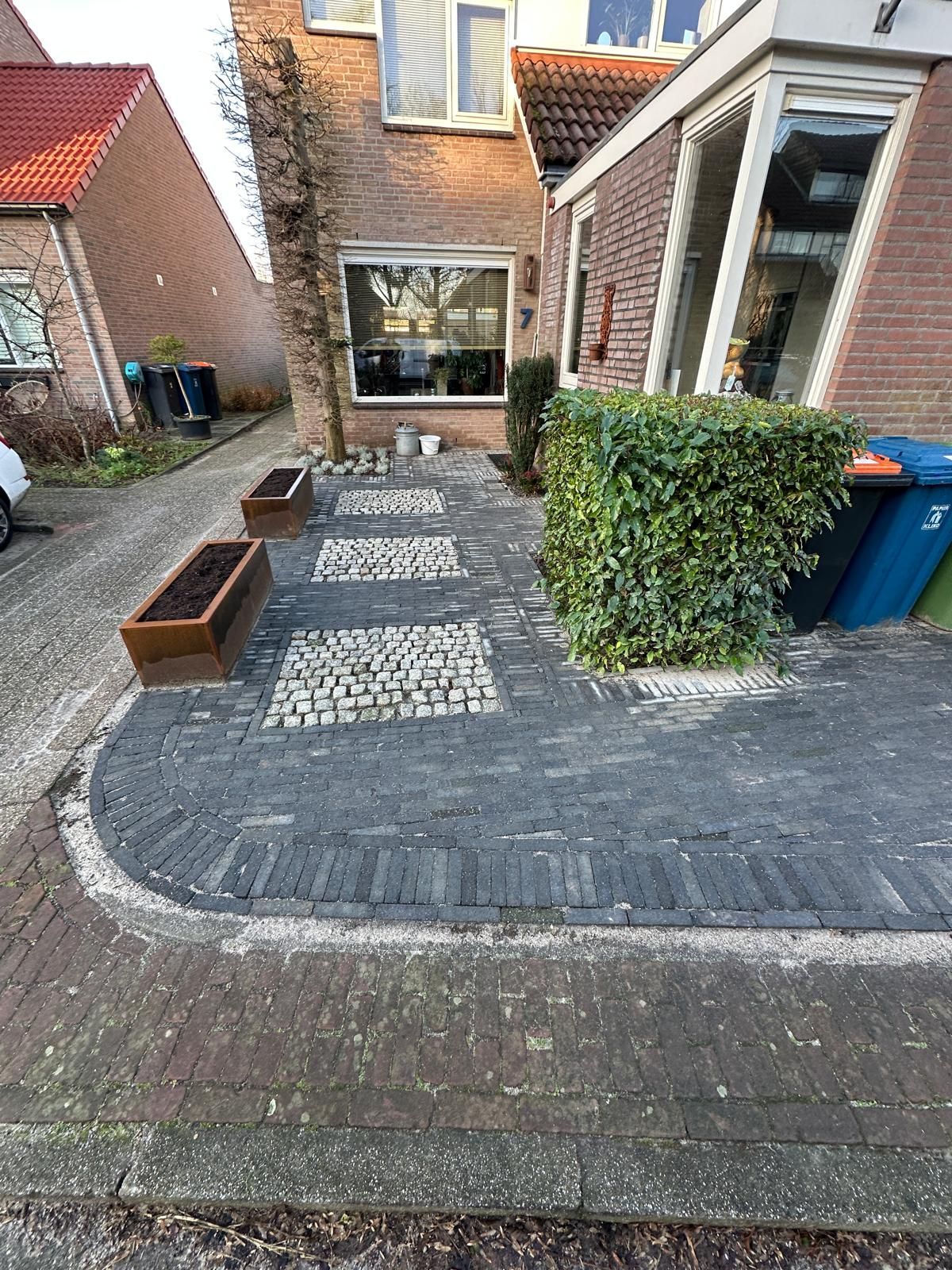 Een pad van grijze bakstenen en kasseien leidt naar een gebouw, geflankeerd door plantenbakken en groene hagen.