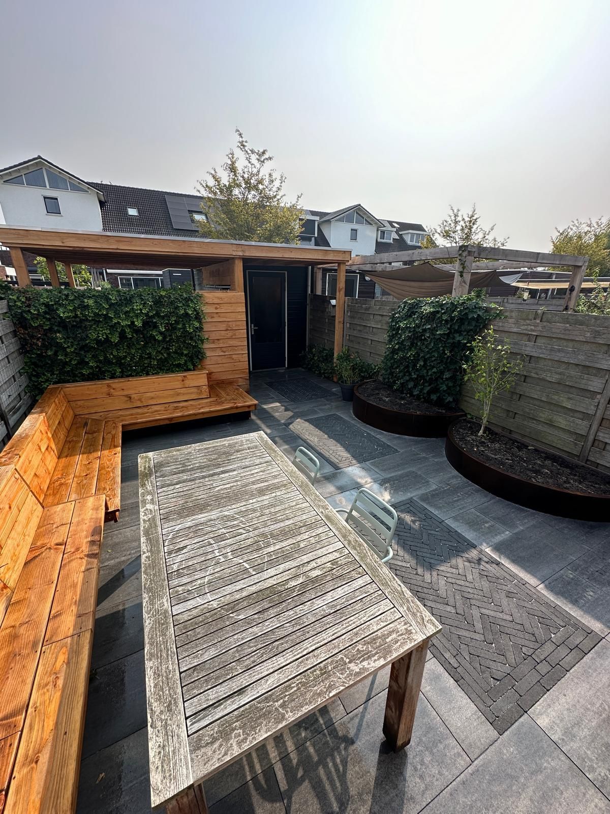 Buitenpatio met houten meubilair, waaronder een tafel en een bank, omgeven door groen en een schutting.