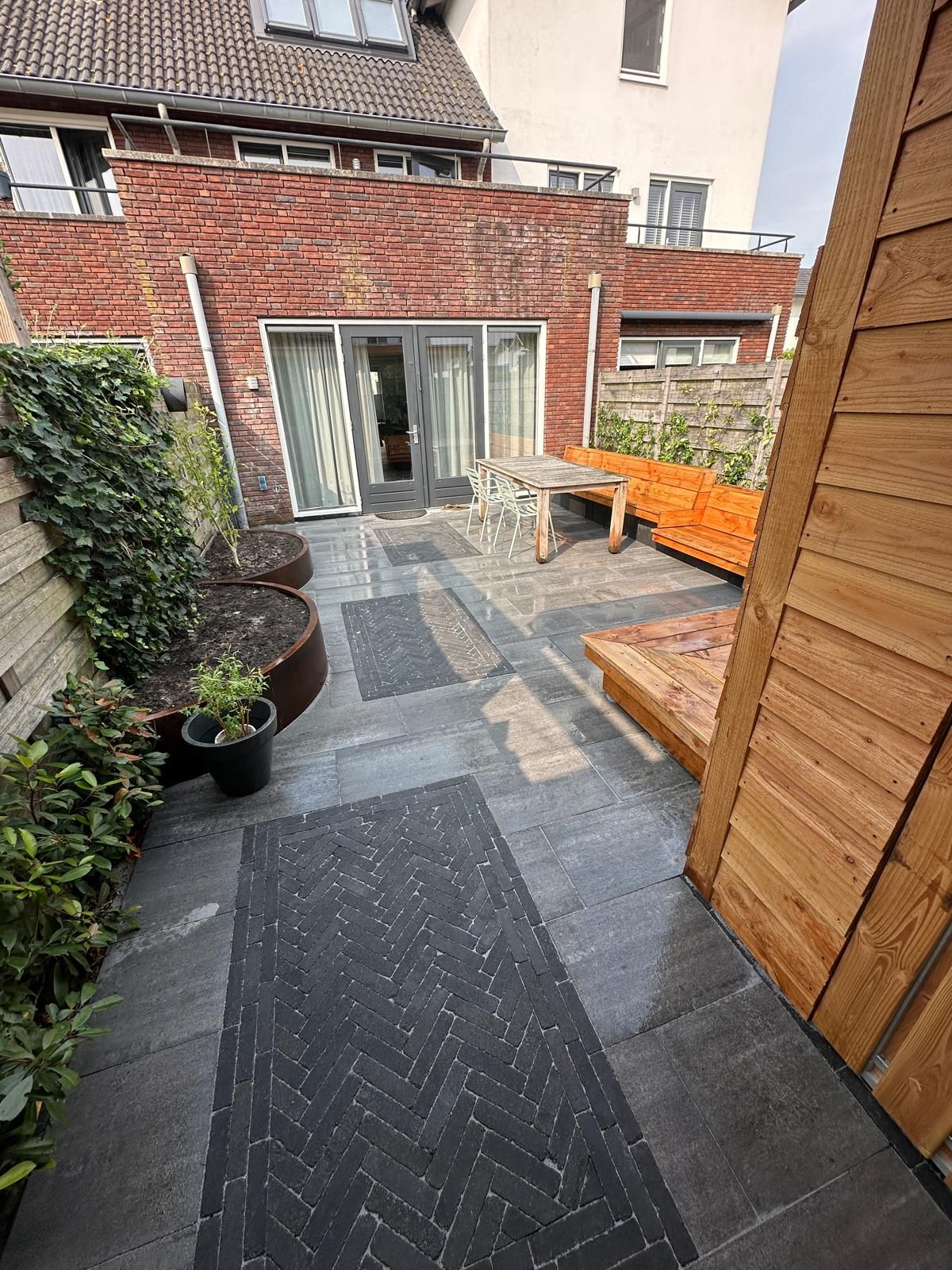 Terras in de achtertuin met een stenen bijgebouw, donkergrijze bestrating, houten plantenbakken en tuinmeubilair.