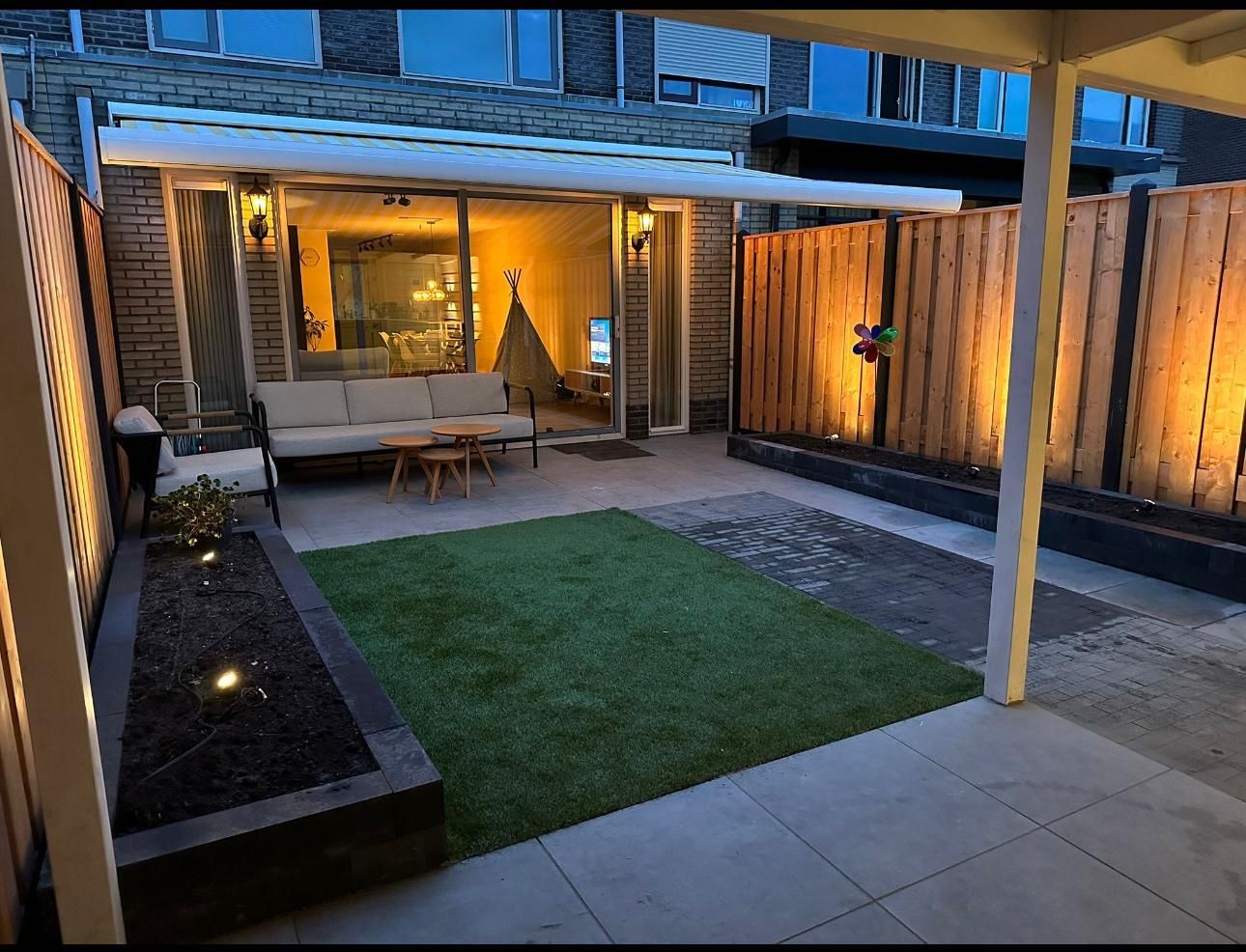 Gezellig terras in de achtertuin 's avonds met een bank, houten schutting en sfeervolle verlichting.