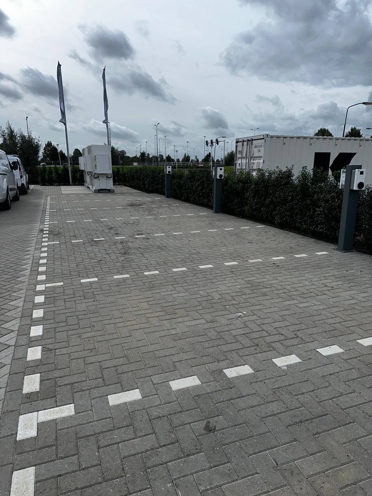 Parkeerplaats met bakstenen bestrating, witte stippellijnen en laadstations. Groene hagen omzomen de achterkant.