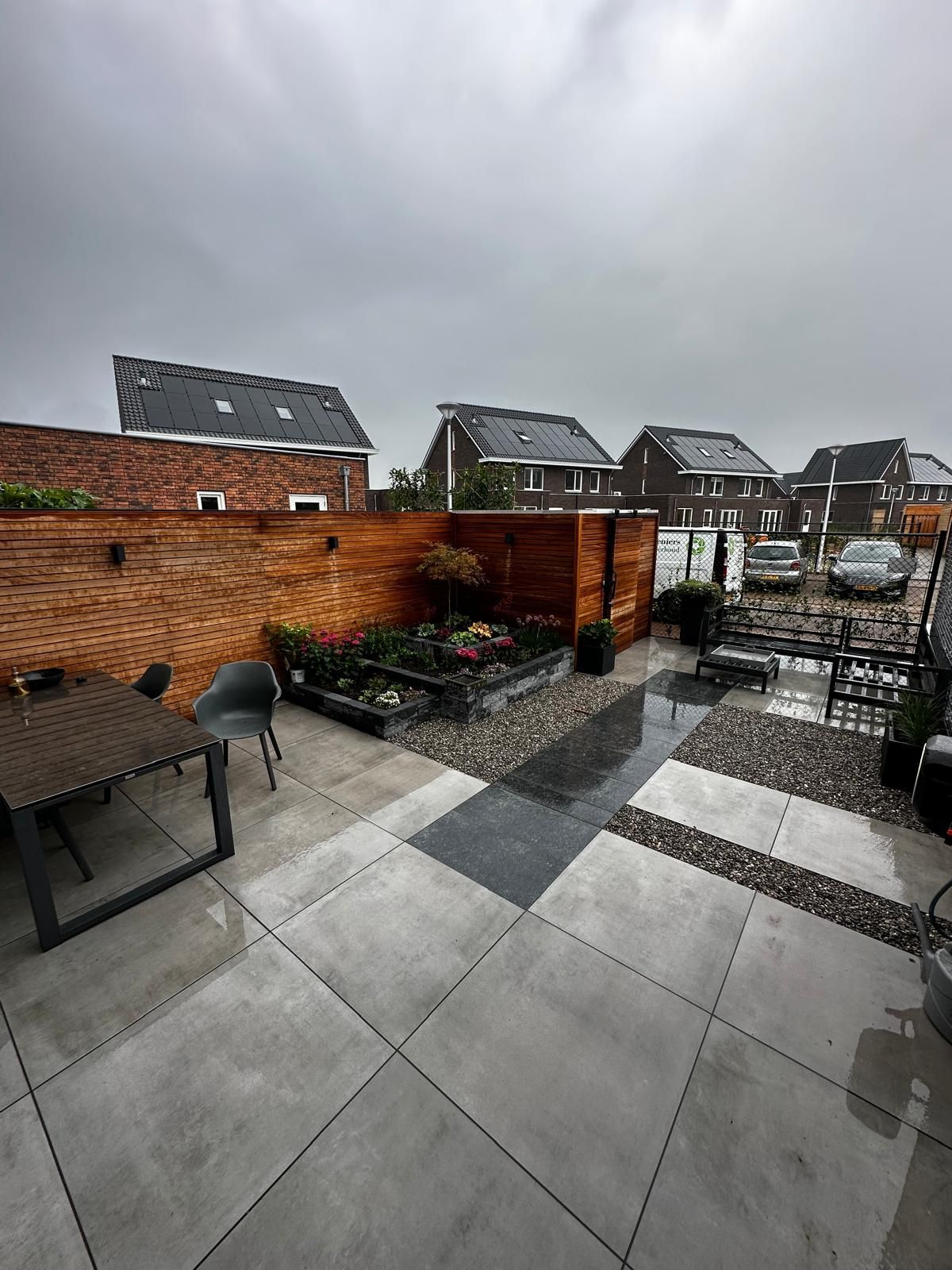 Terras in de achtertuin met grijze bestrating, bakstenen muur, houten schutting en een donkere hemel.