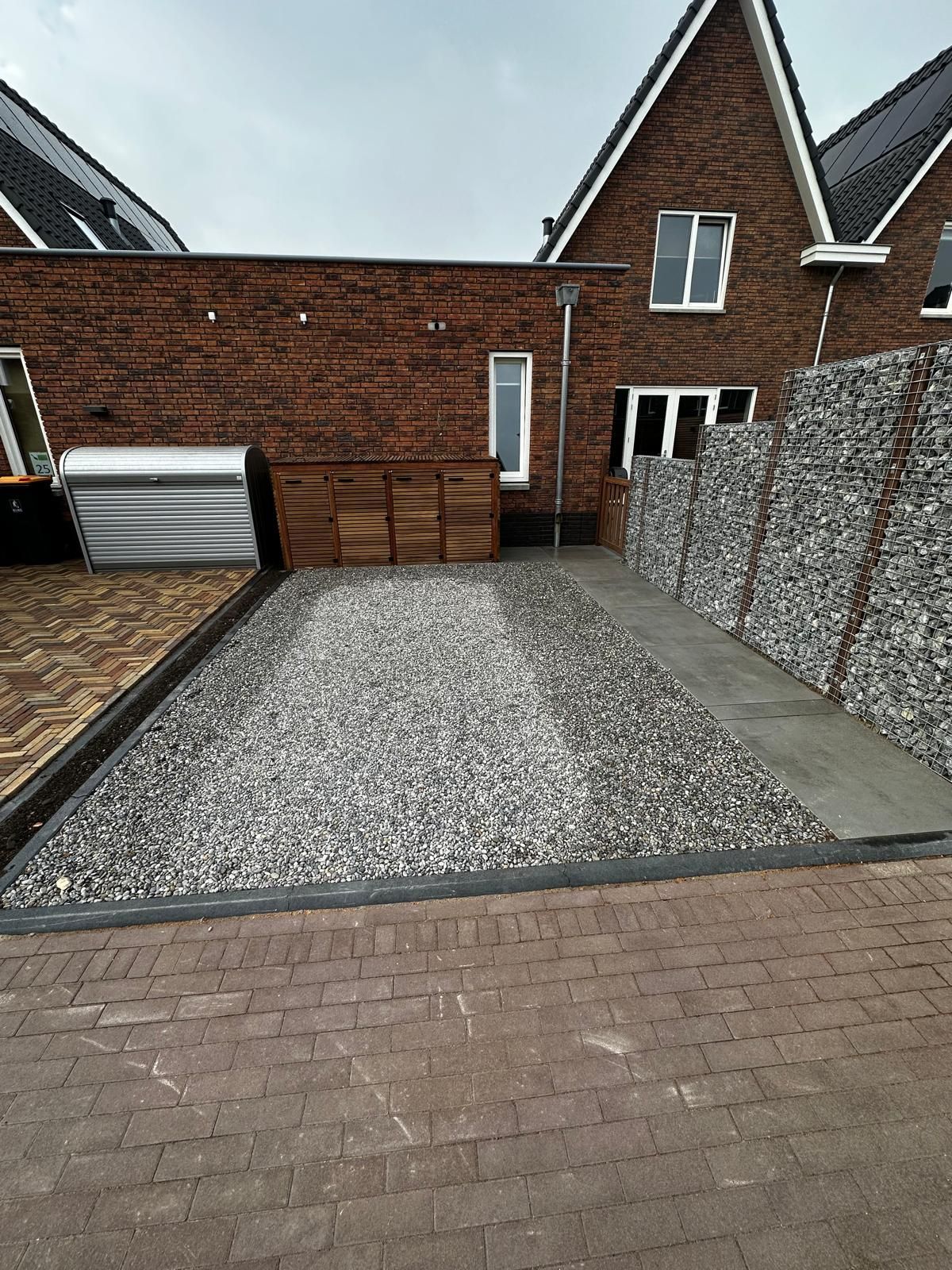 Een grindpad, omzoomd met een zwarte rand, grenst aan een keermuur van gestapelde stenen en leidt naar een bakstenen huis.