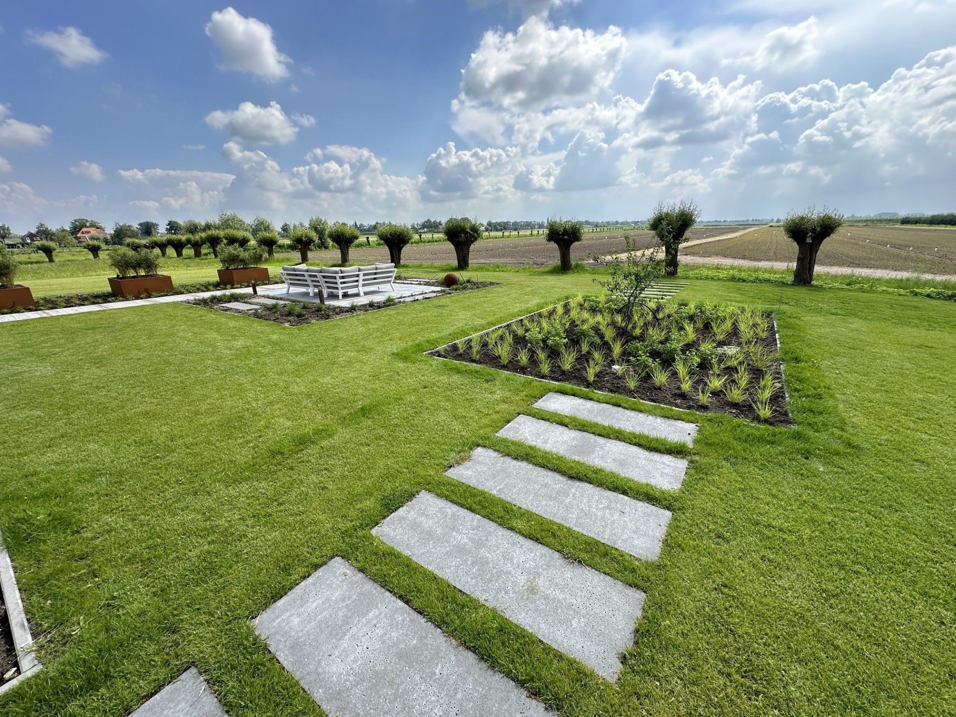 Een keurig onderhouden gazon met een stenen pad, aangelegde beplanting en bomen tegen een bewolkte blauwe hemel.