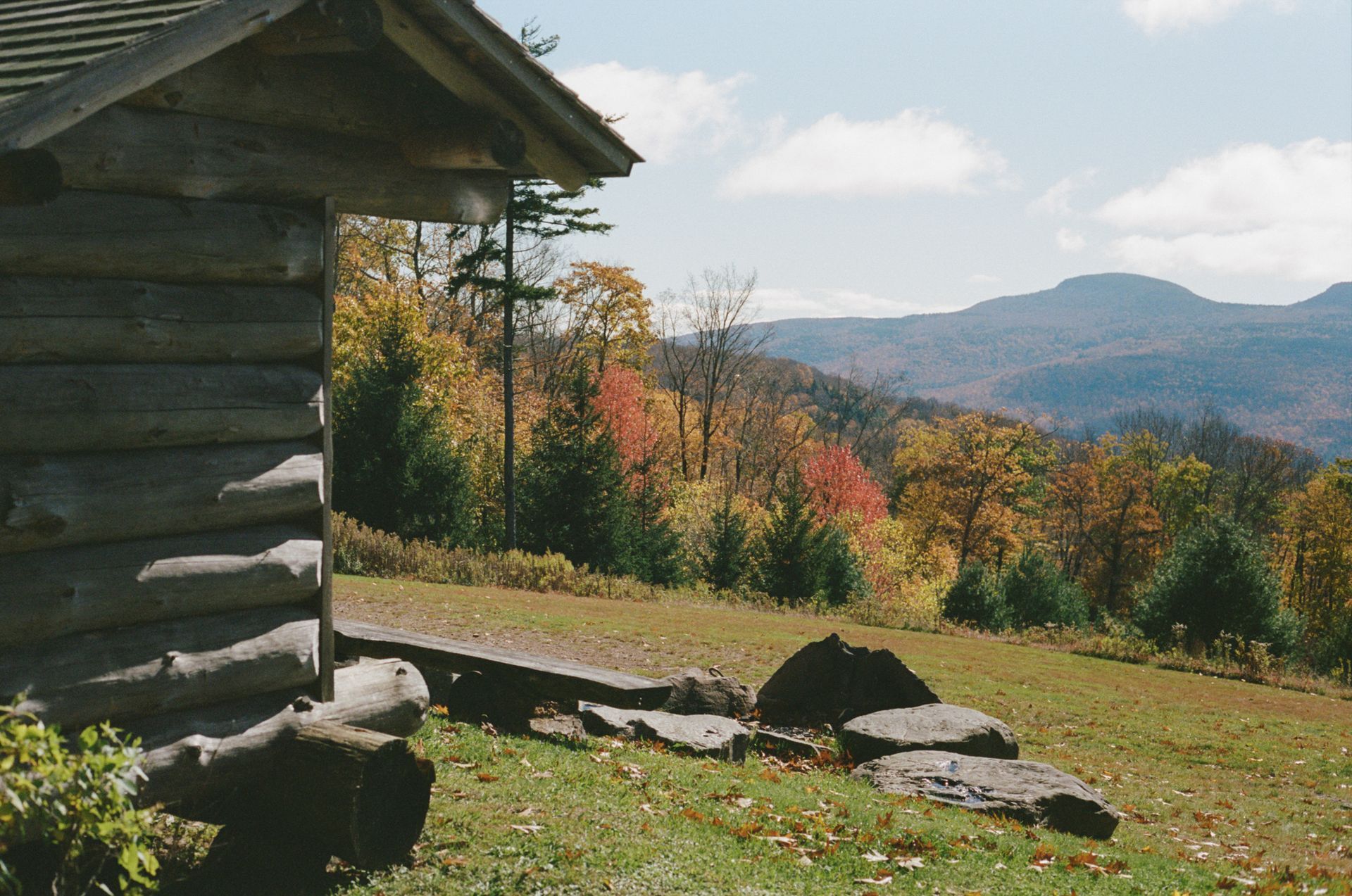 A log cabin sits in the middle of a grassy field with mountains in the background