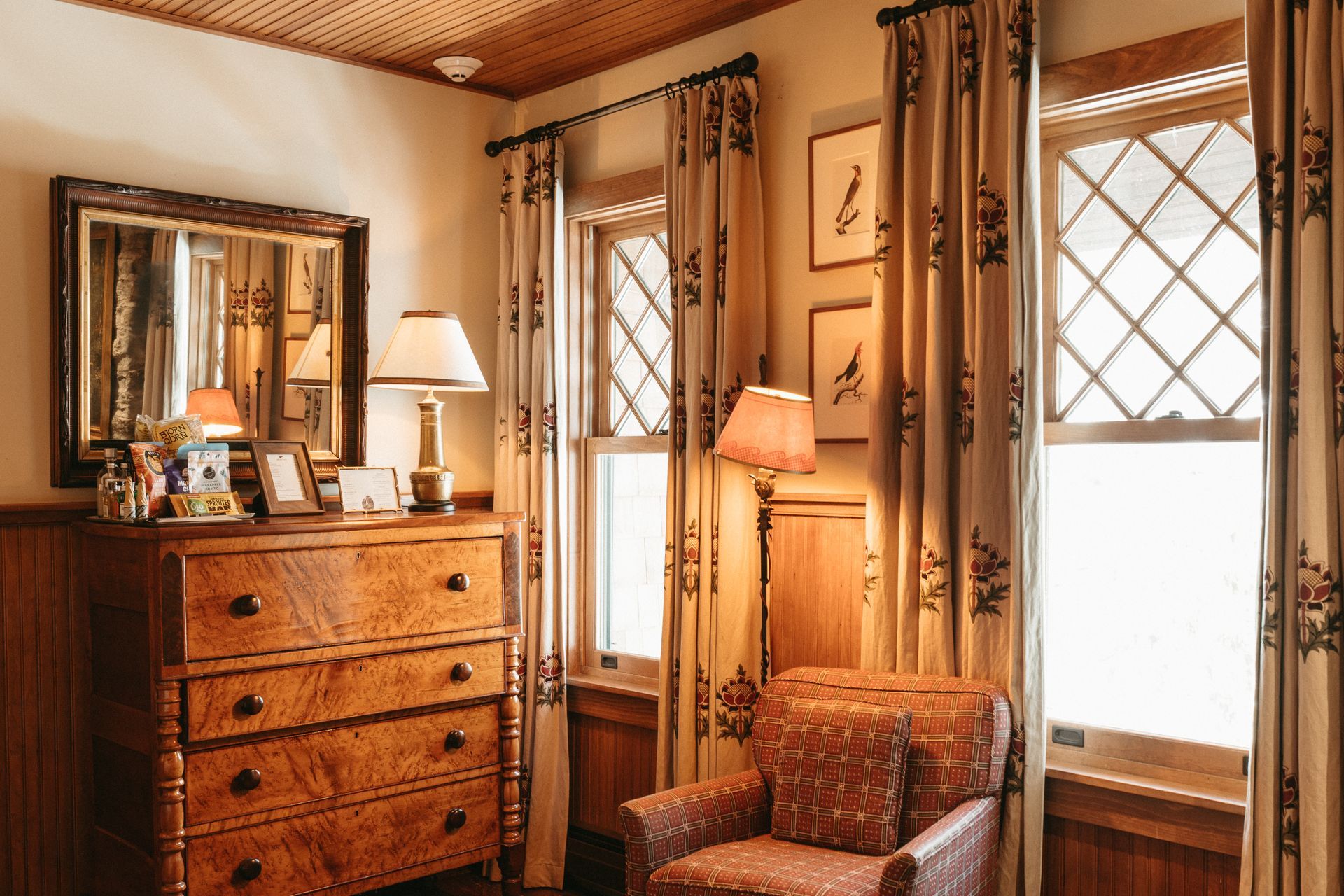 A living room with a dresser , chair , lamp , mirror and curtains.