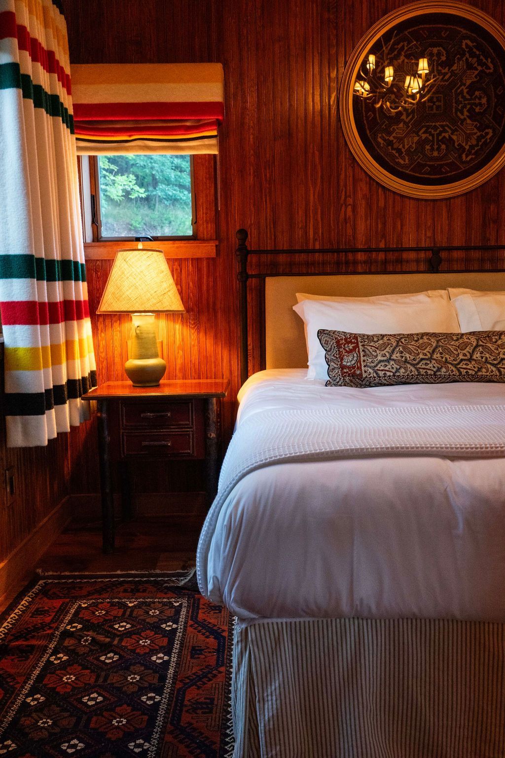 A bedroom with a bed , nightstand , lamp and window.