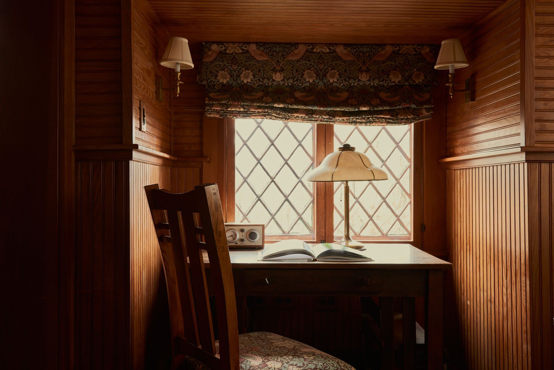 A desk with a lamp and a book on it in front of a window.