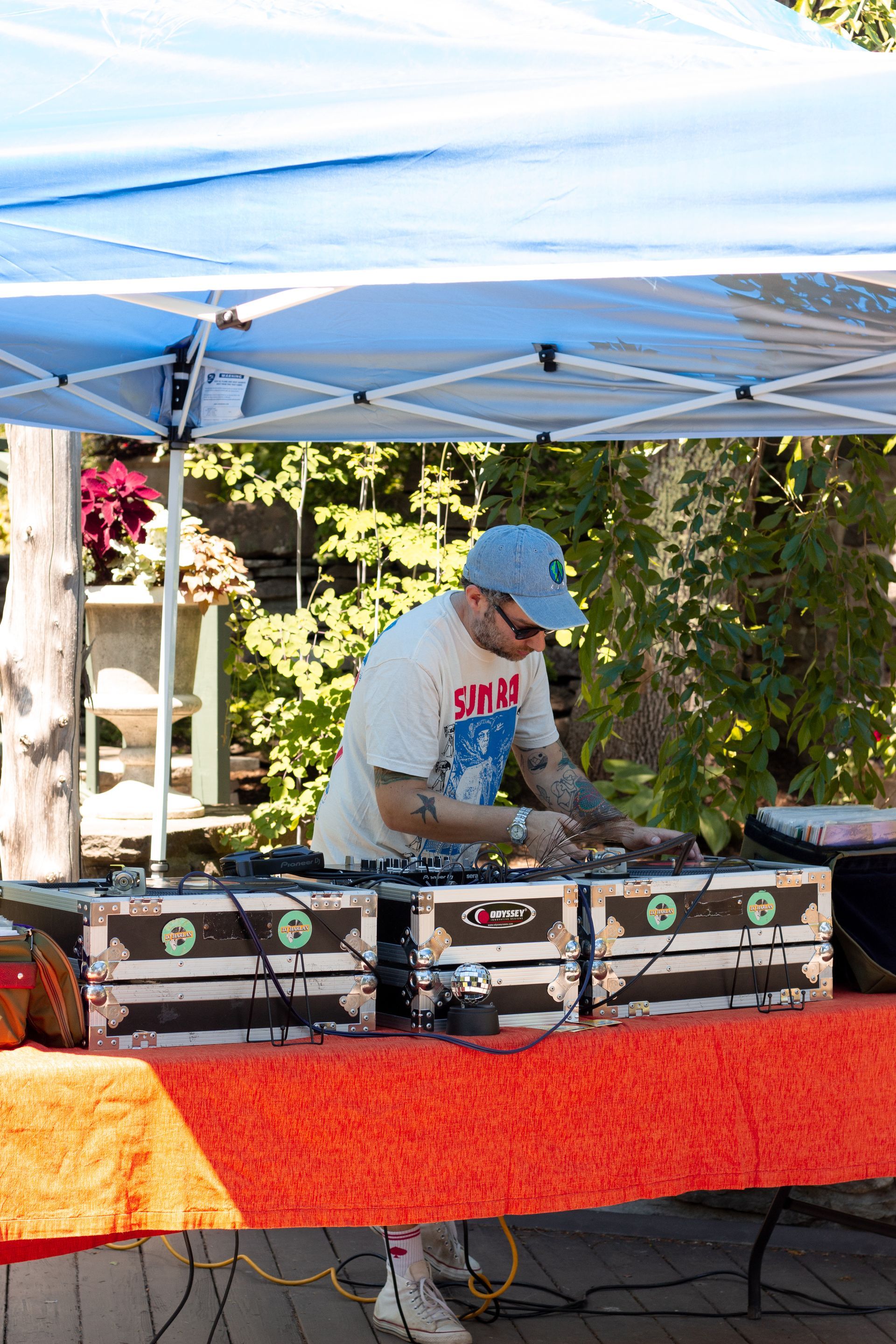 DJ performing outdoors under a blue canopy behind a red table