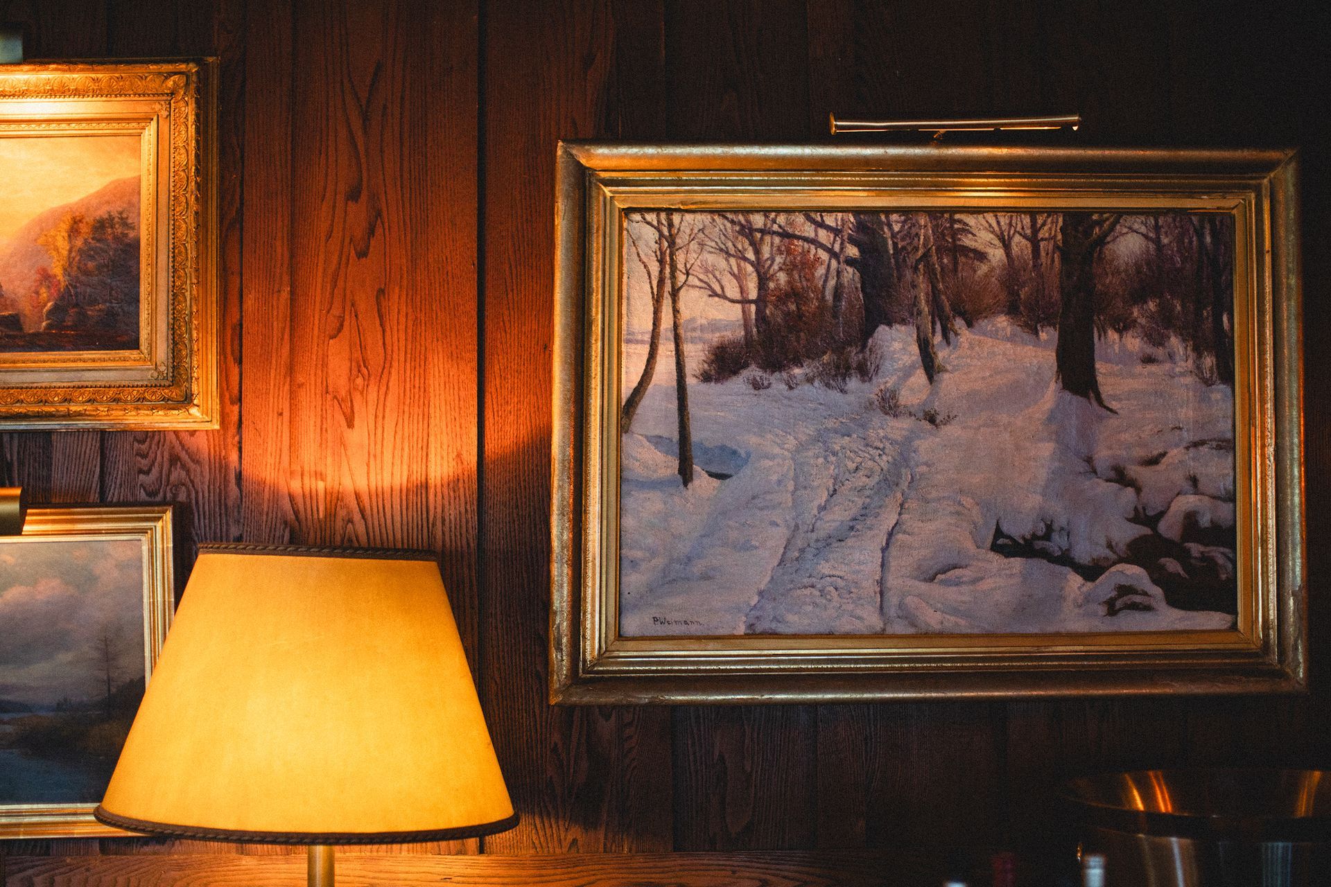 A painting of a snowy forest is hanging on a wooden wall next to a lamp.