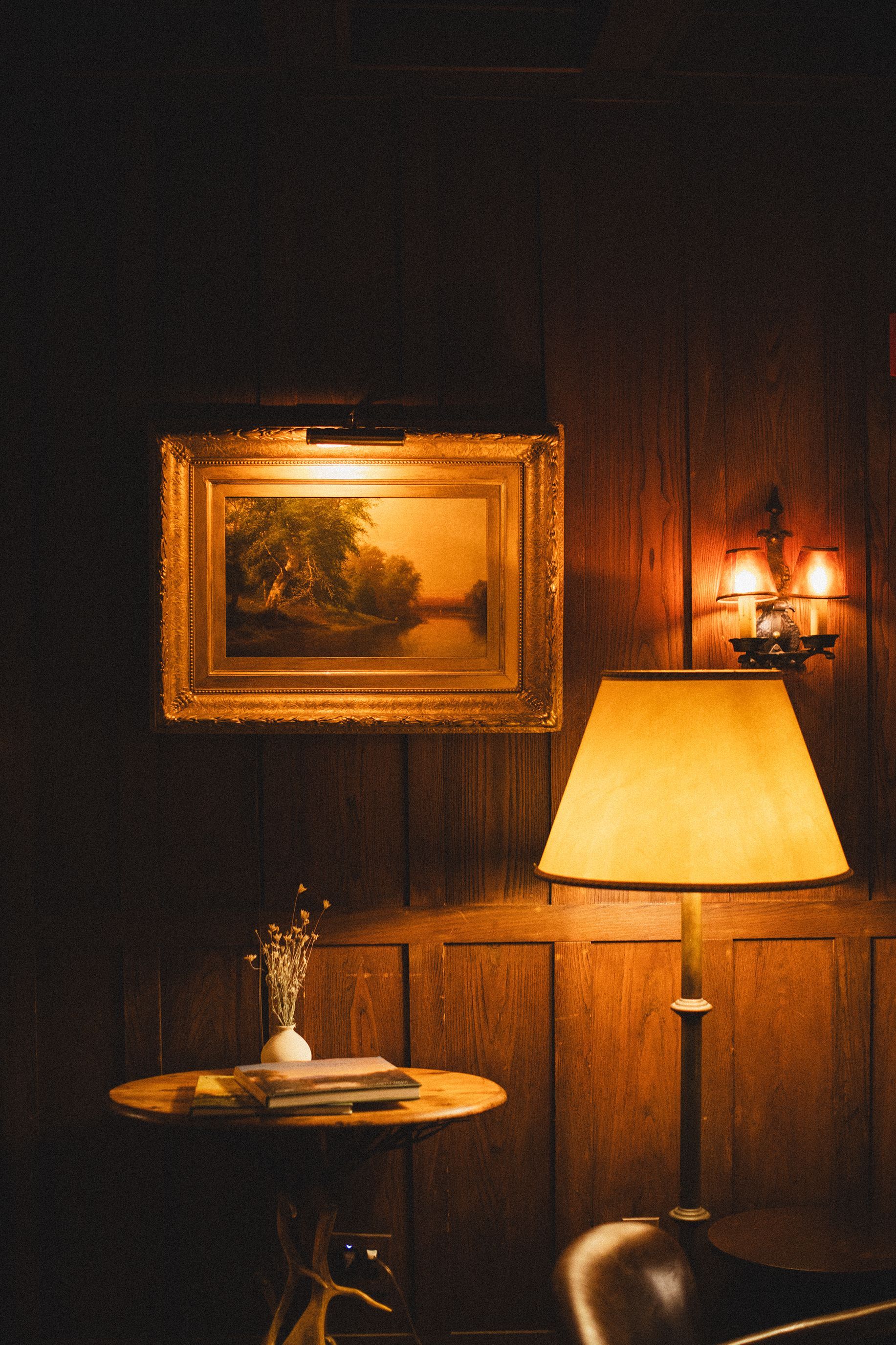 A lamp is lit up in a dark room with a painting on the wall above it.