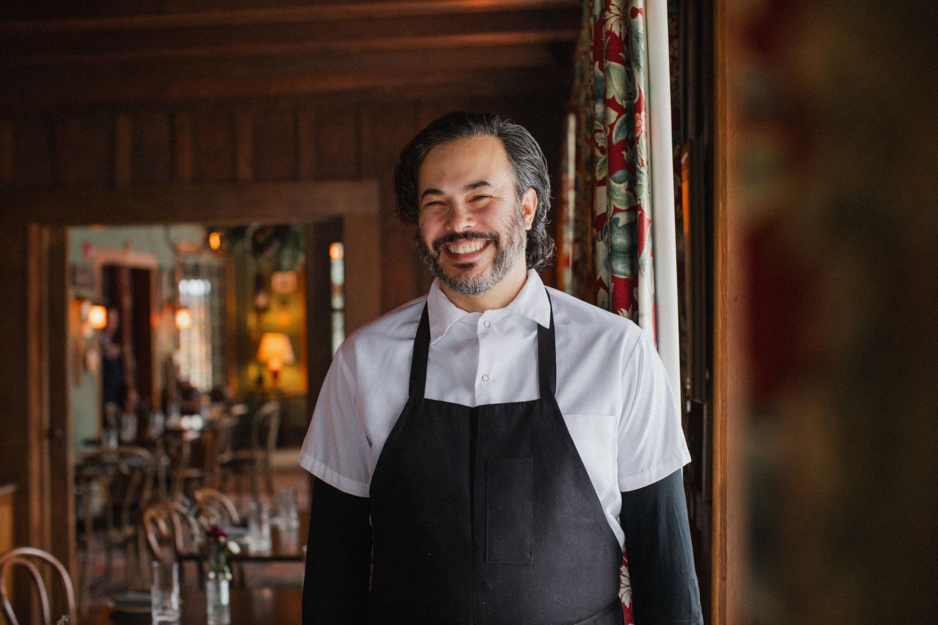 A man with a beard is wearing an apron and smiling in a restaurant.