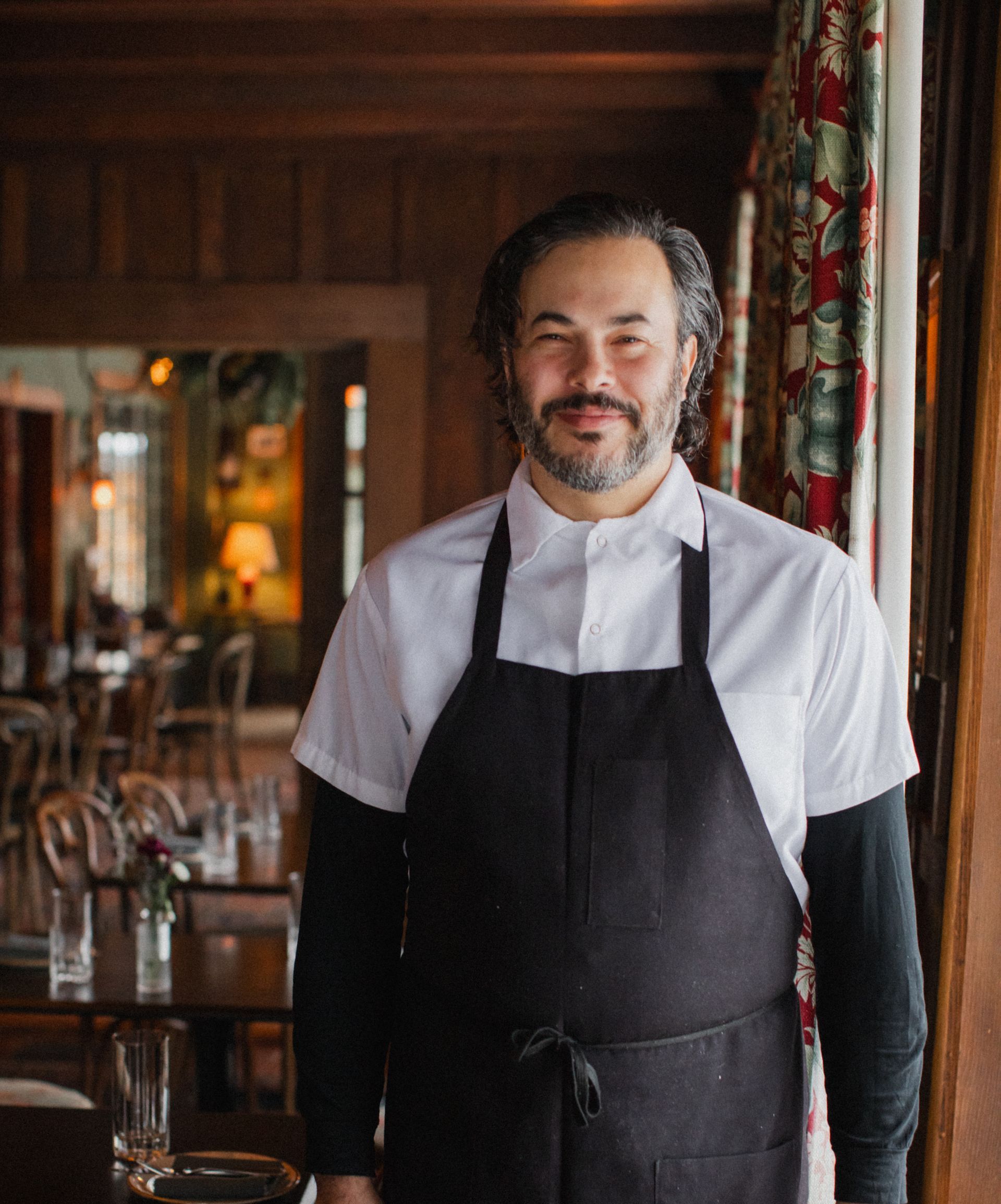 A man wearing an apron and a white shirt is standing in a restaurant.
