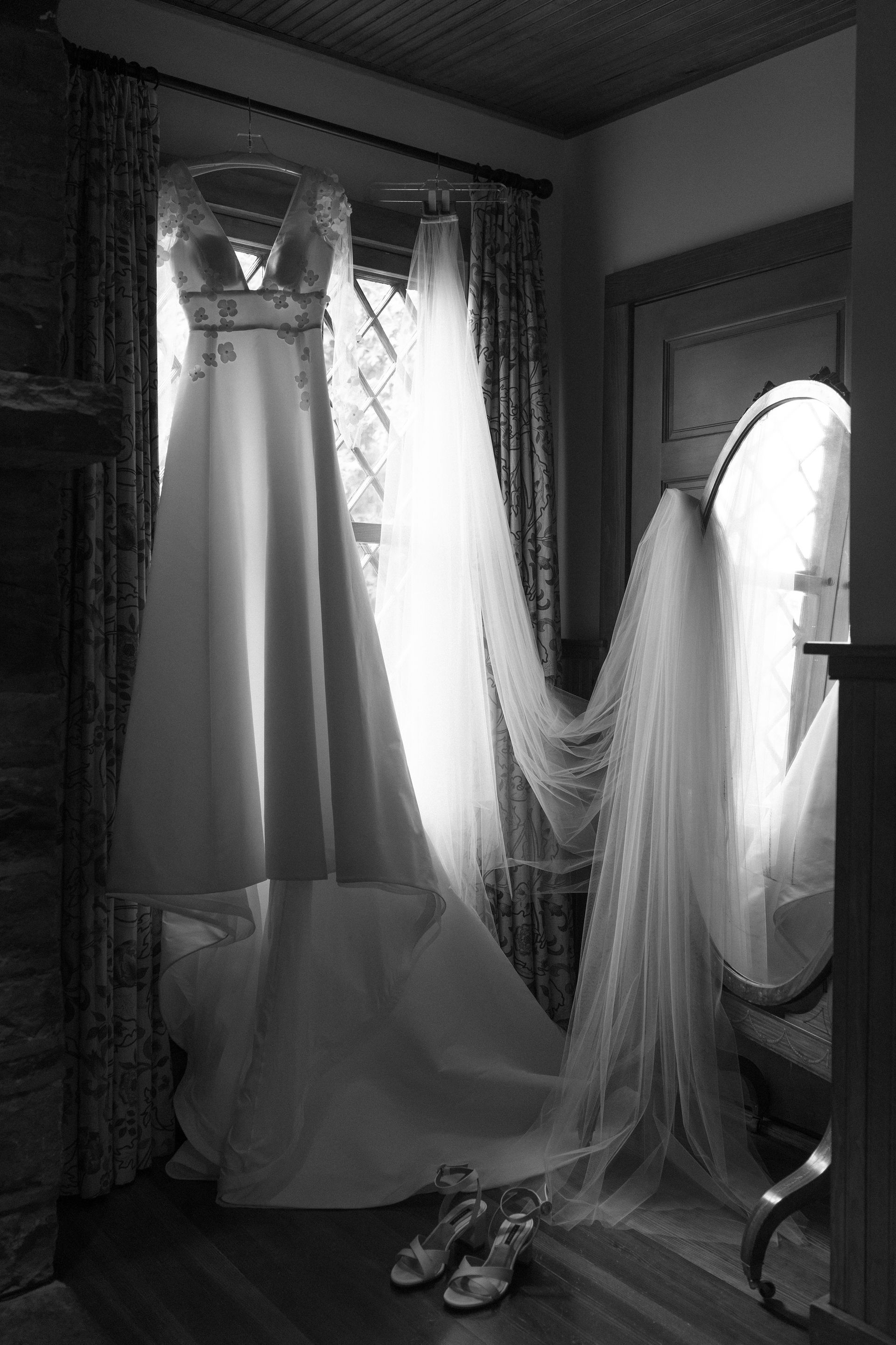 A black and white photo of a wedding dress and veil hanging on a window.