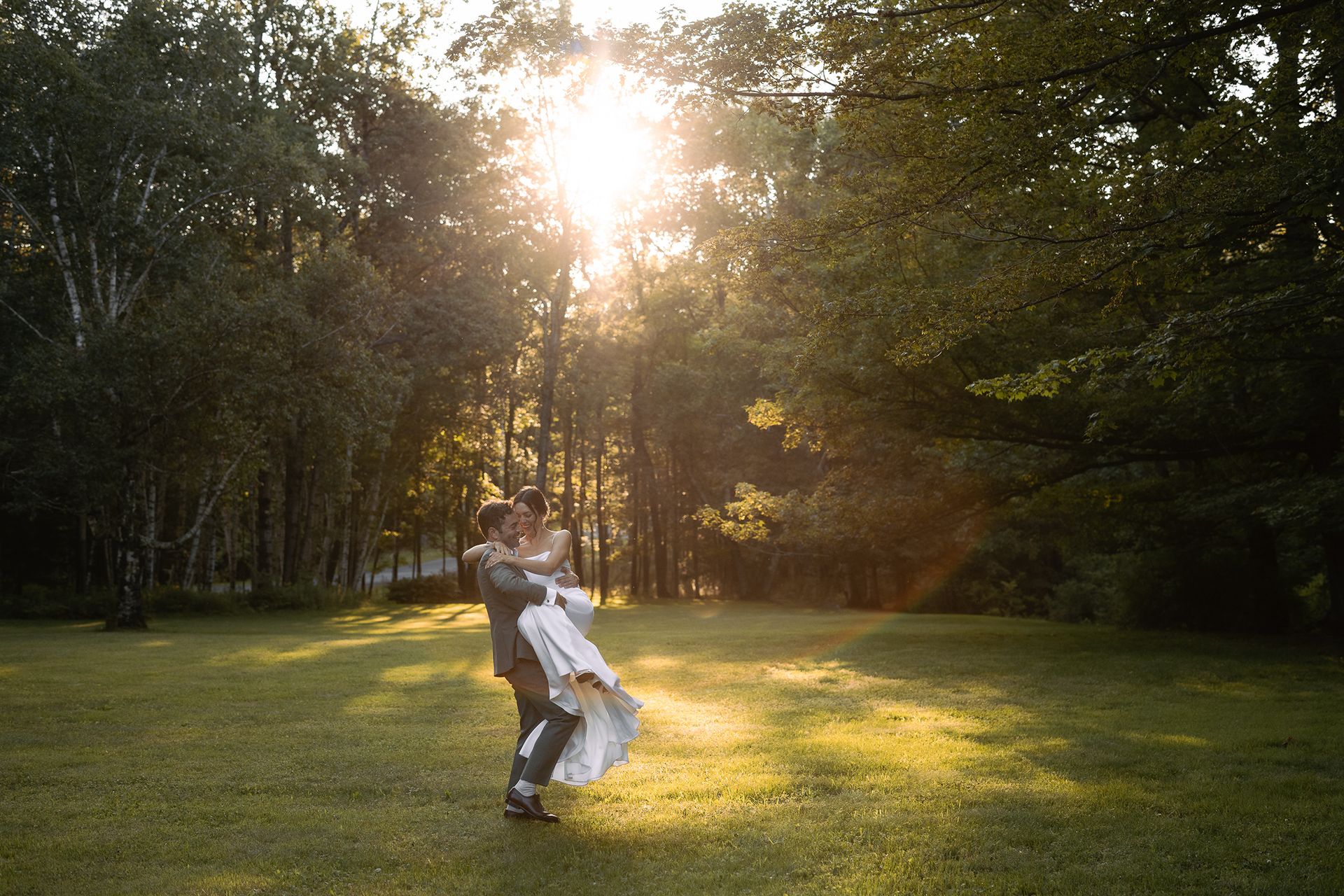 A bride and groom are dancing in a field with the sun shining through the trees.