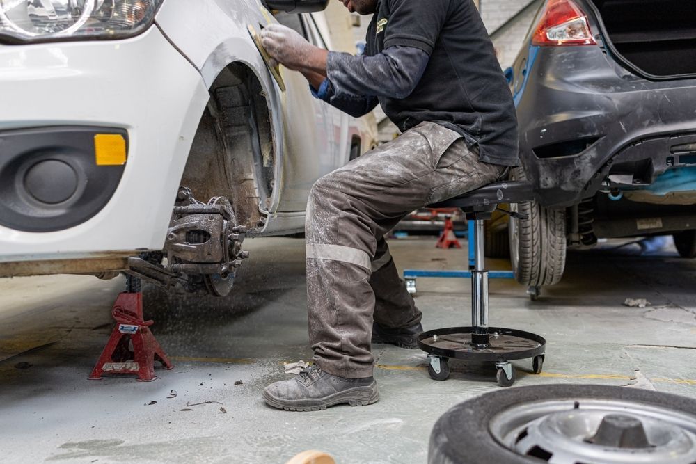 A Man is Sitting on a Stool Working on a Car in a Garage — C & C Panel & Paint Mackay In Mackay, QLD