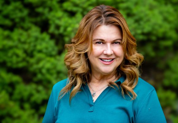 Smiling woman with shoulder length brown hair in a teal top against a blurred green outdoor background
