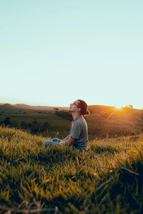 Young man sitting in tall golden grass at sunset, with sunlit hills in the background. He has dark hair in a bun and is wearing glasses, a grey tee shirt, and jeans.