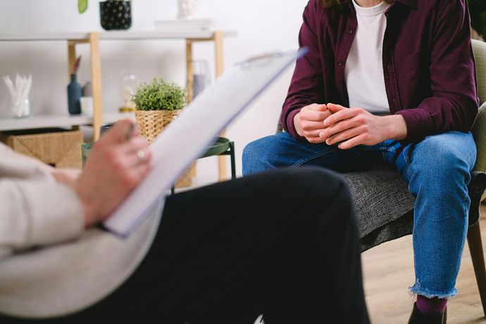 Therapist taking notes while a seated client in jeans and a maroon shirt talks in a counseling office.