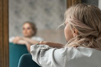 Woman resting her arms on a chair, seen from behind, facing herself in a mirror in a softly lit room
