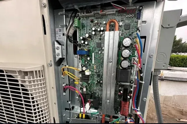 Open electrical control panel with circuit board, wires, and capacitors on an outdoor HVAC unit