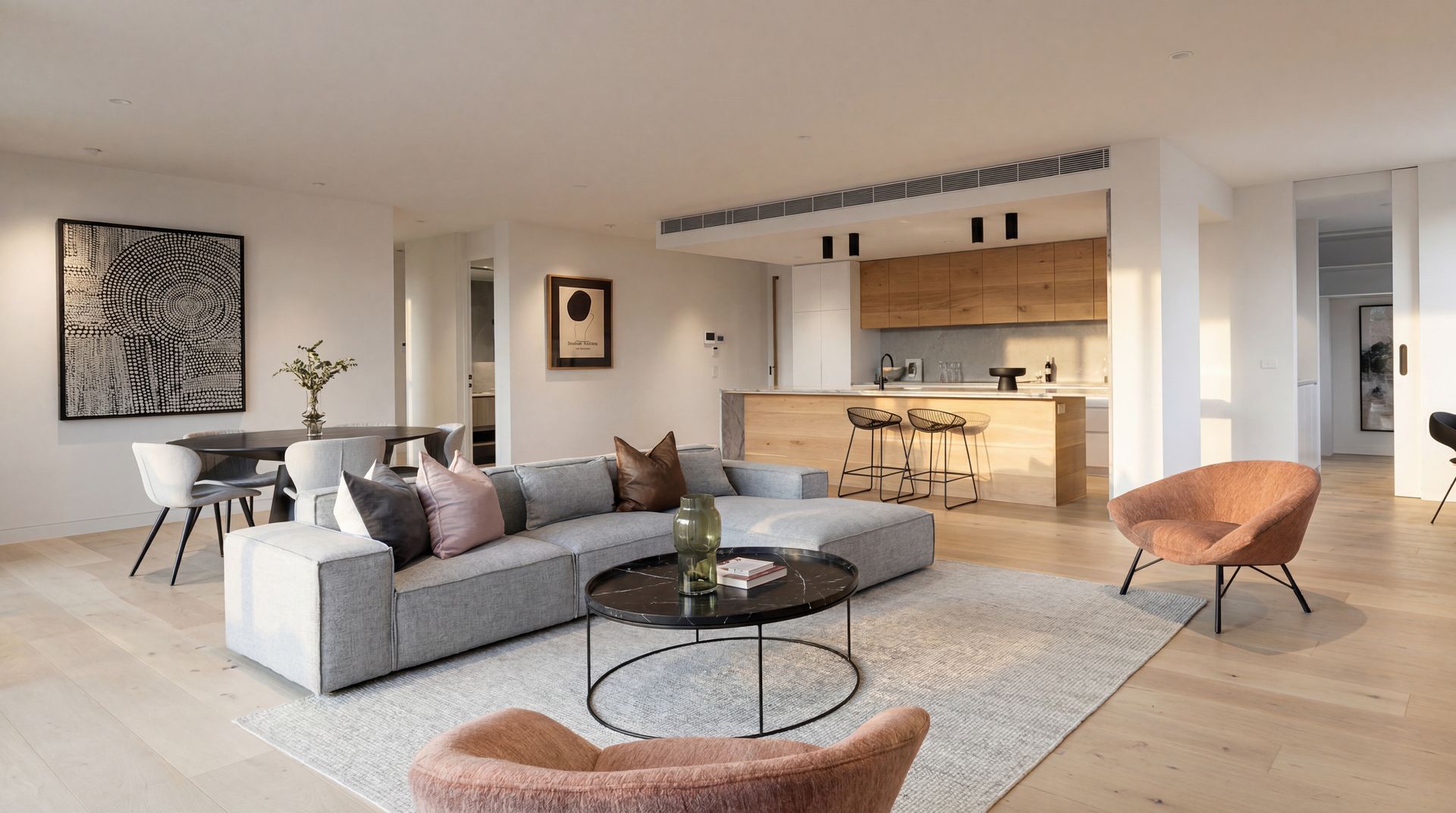 Modern open-plan living room with gray sectional, round coffee table, dining area, and warm wood kitchen.