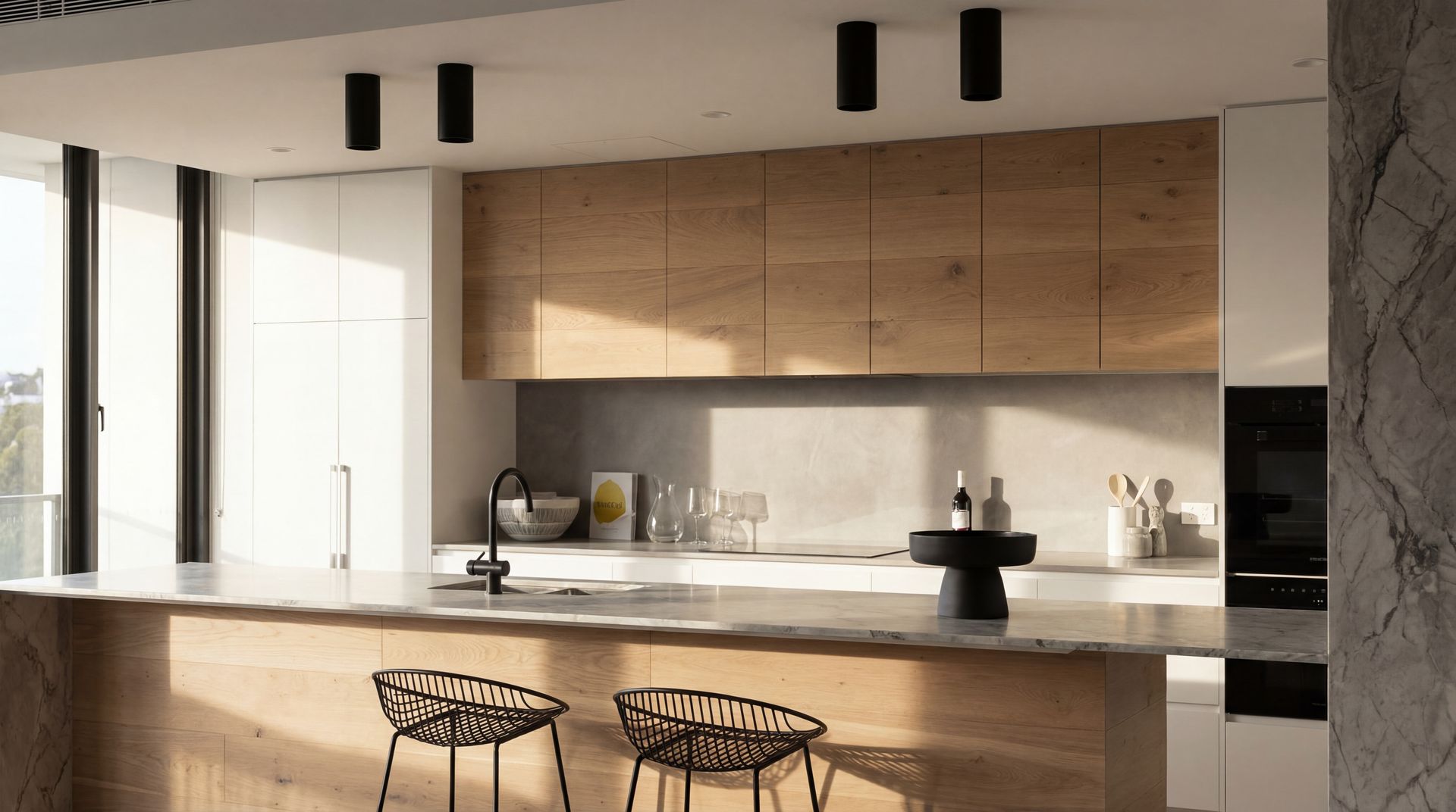 Modern kitchen with wood cabinets, white countertop, black stools, and large windows in warm natural light