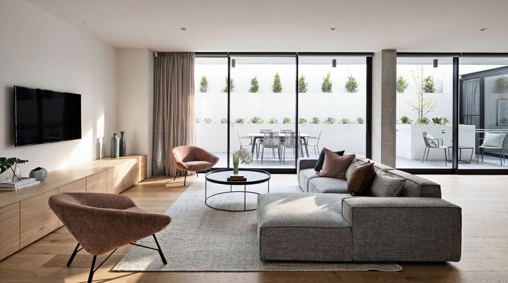 Modern living room with gray sofa, pink chairs, coffee table, and floor-to-ceiling windows opening to a patio.