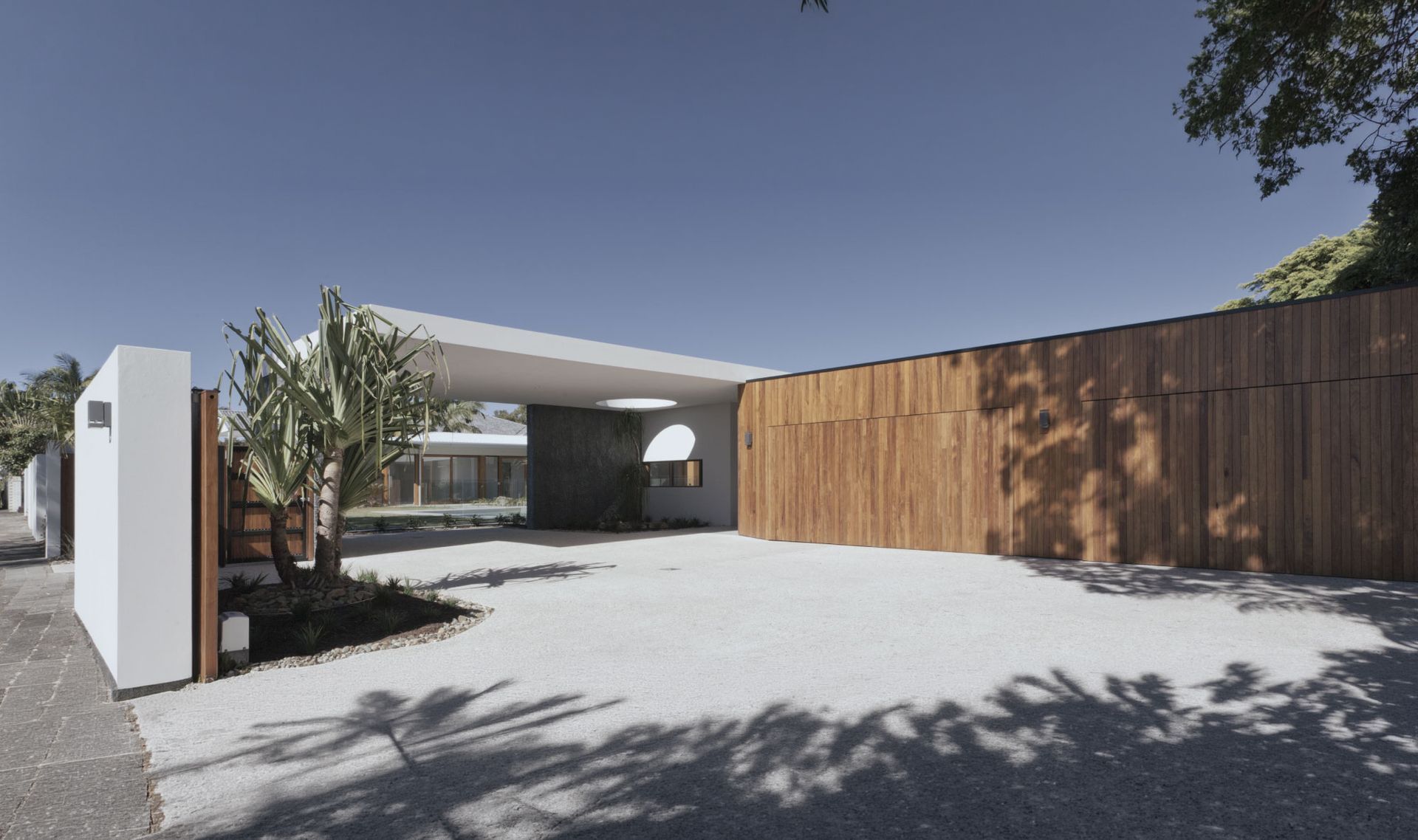 Modern home entrance with white walls, wooden fence, and gravel driveway under a clear blue sky