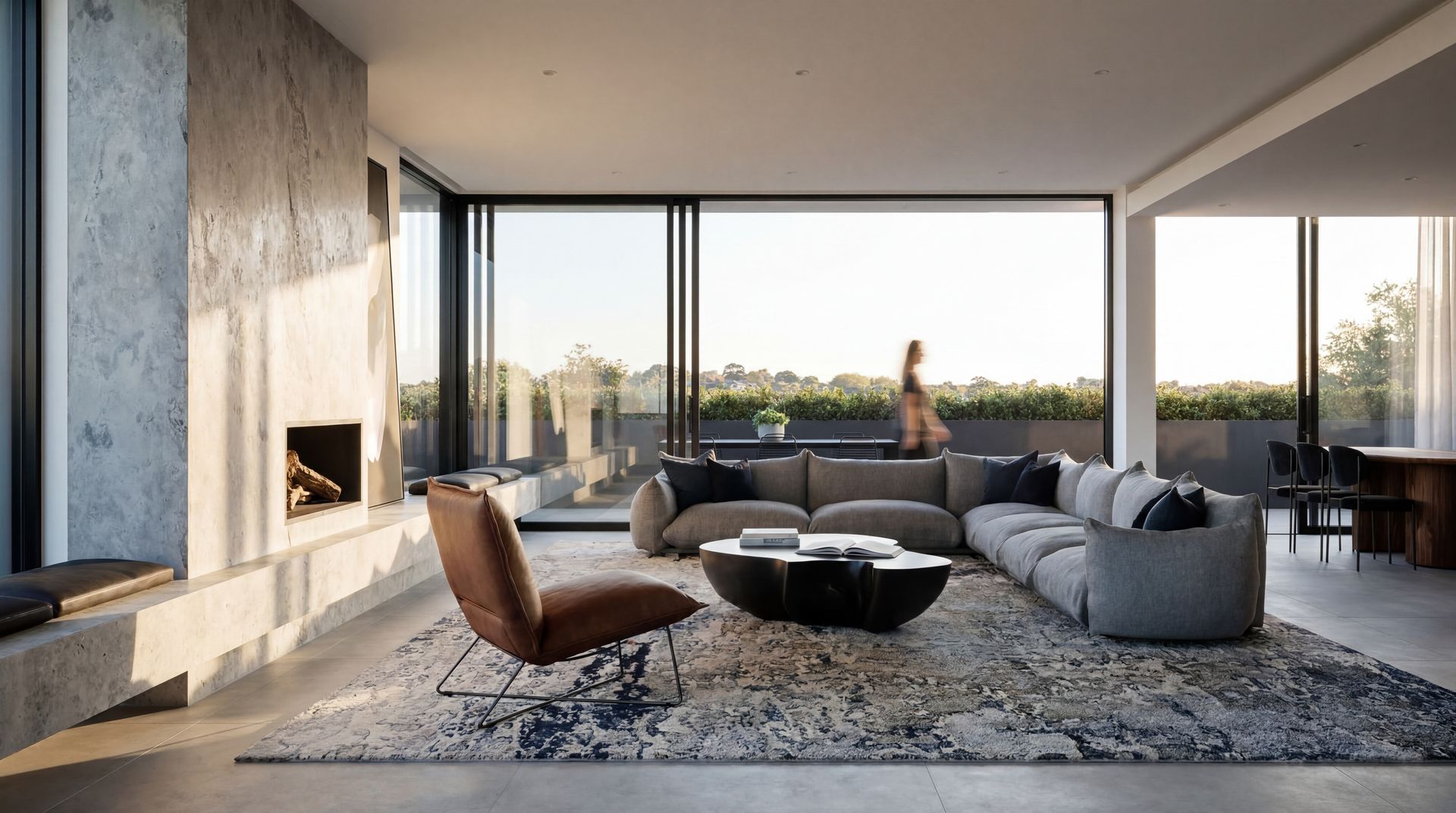 Modern living room with floor-to-ceiling windows, gray sectional, brown chair, and fireplace overlooking a sunset view
