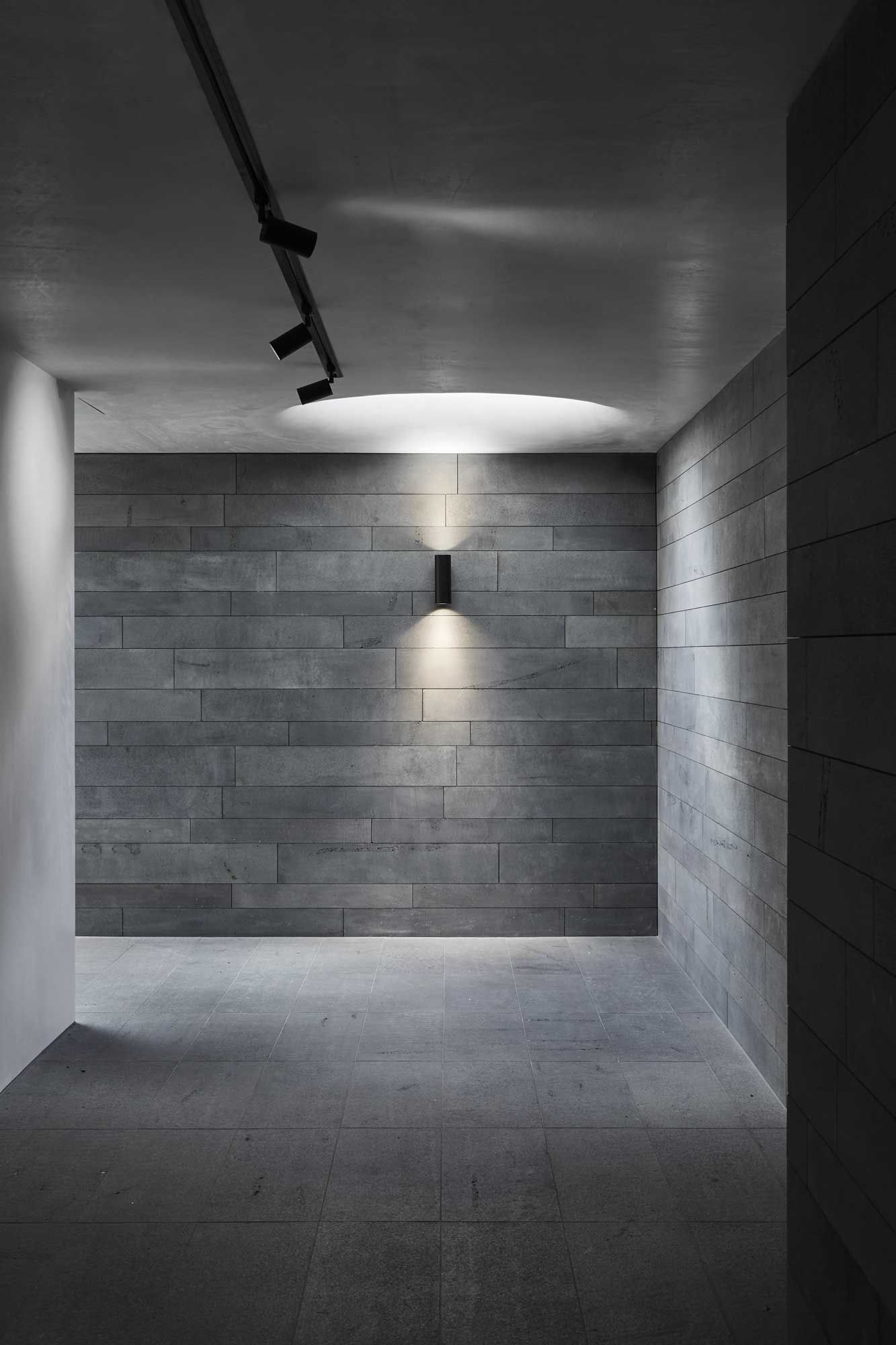 Empty modern hallway with gray stone walls, concrete floor, and a single wall sconce lighting the end wall