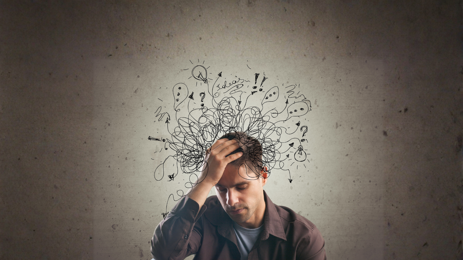 A stressed man holds his head in front of a wall with chaotic scribble-like drawings above him.